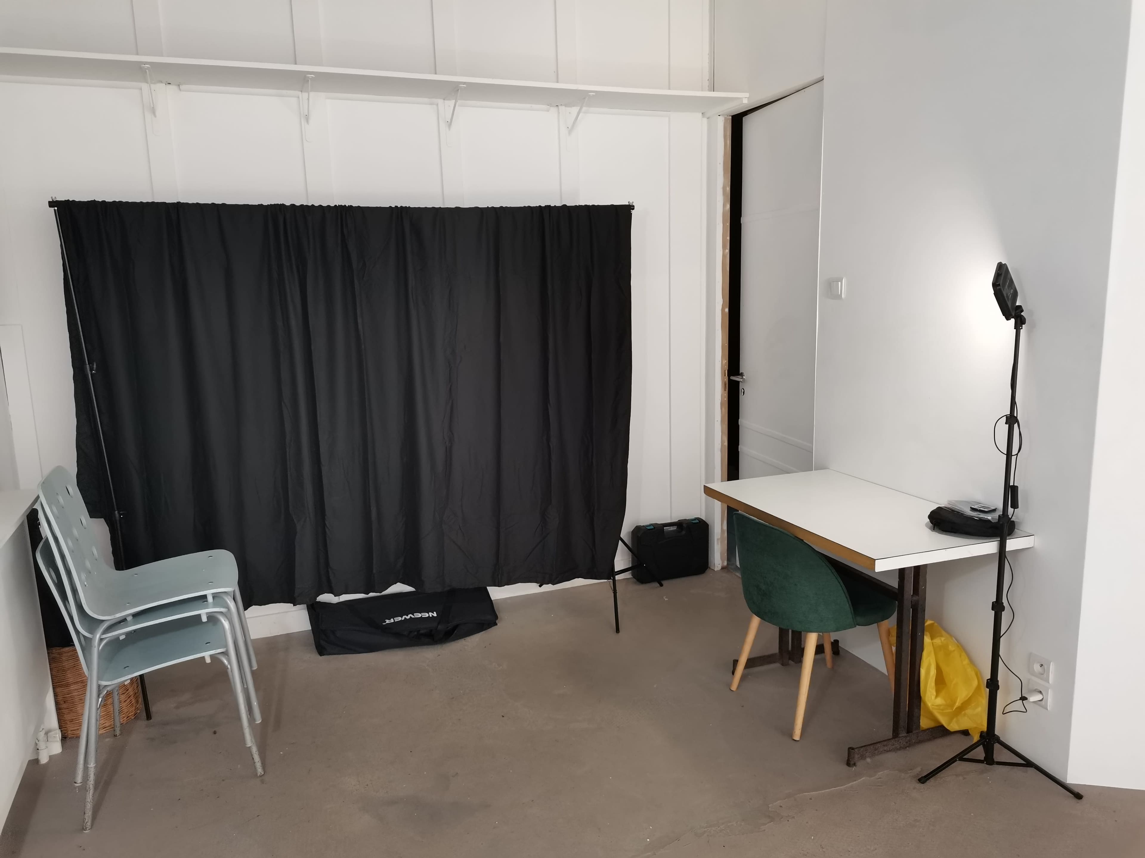A minimalist room with a black curtain, a desk with a lamp, a green chair, and three stacked chairs in the corner.
