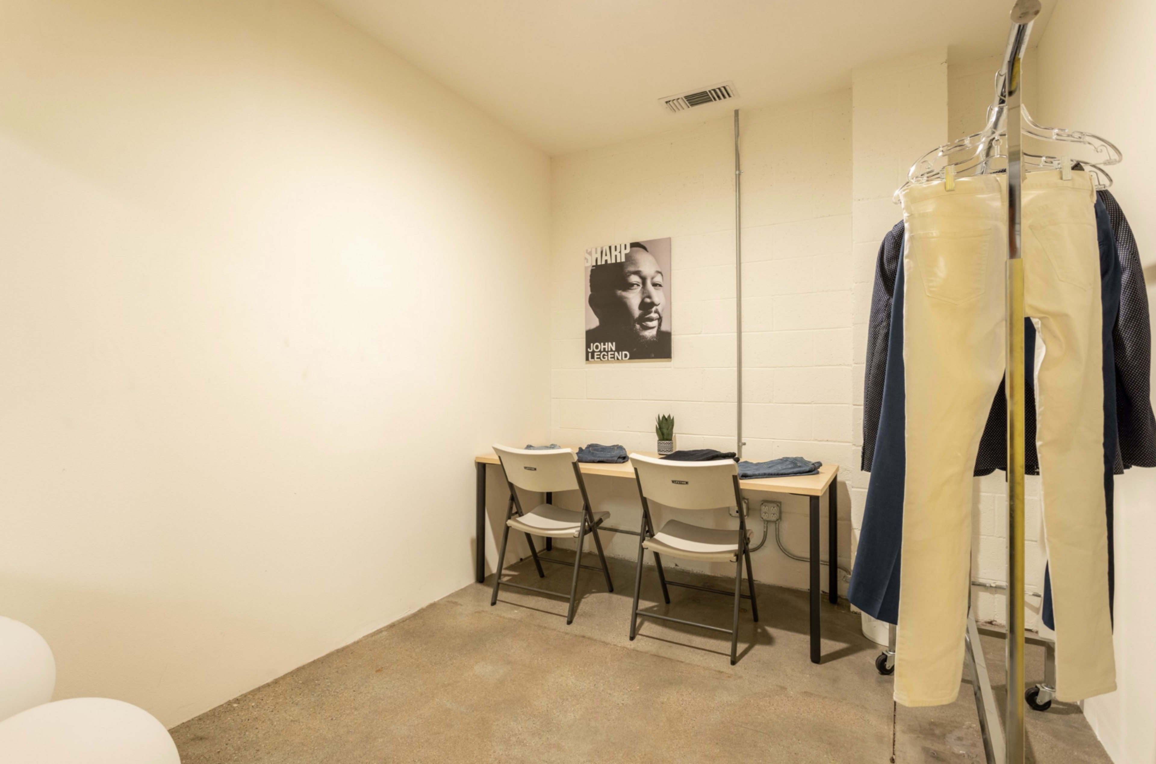 A minimalist room with a table and two chairs, a plant on the table, and hanging clothing on a clothing rack, accompanied by a framed black-and-white photo on the wall.