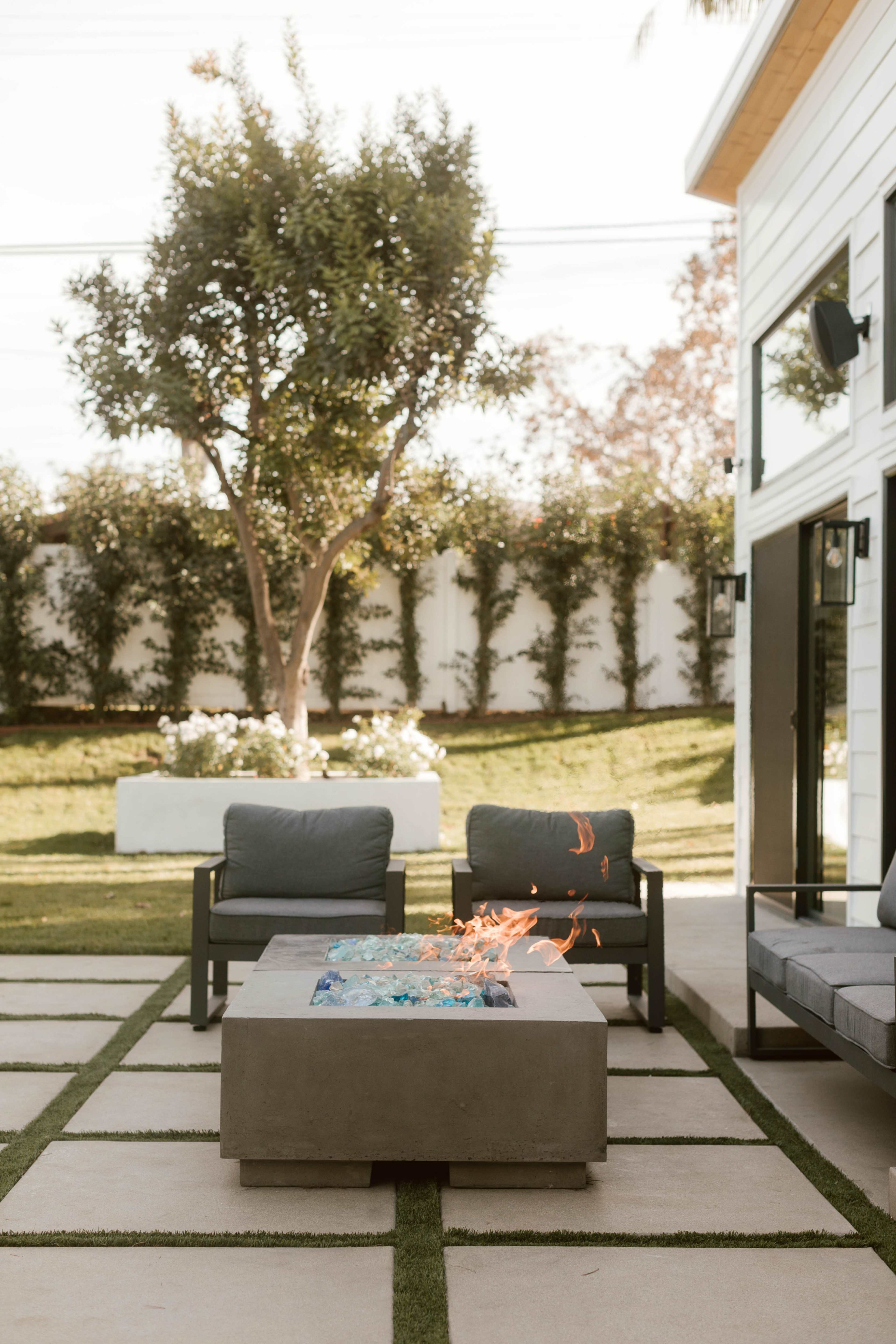 A modern outdoor seating area features a fire pit surrounded by two chairs, with a grassy lawn and trees in the background.