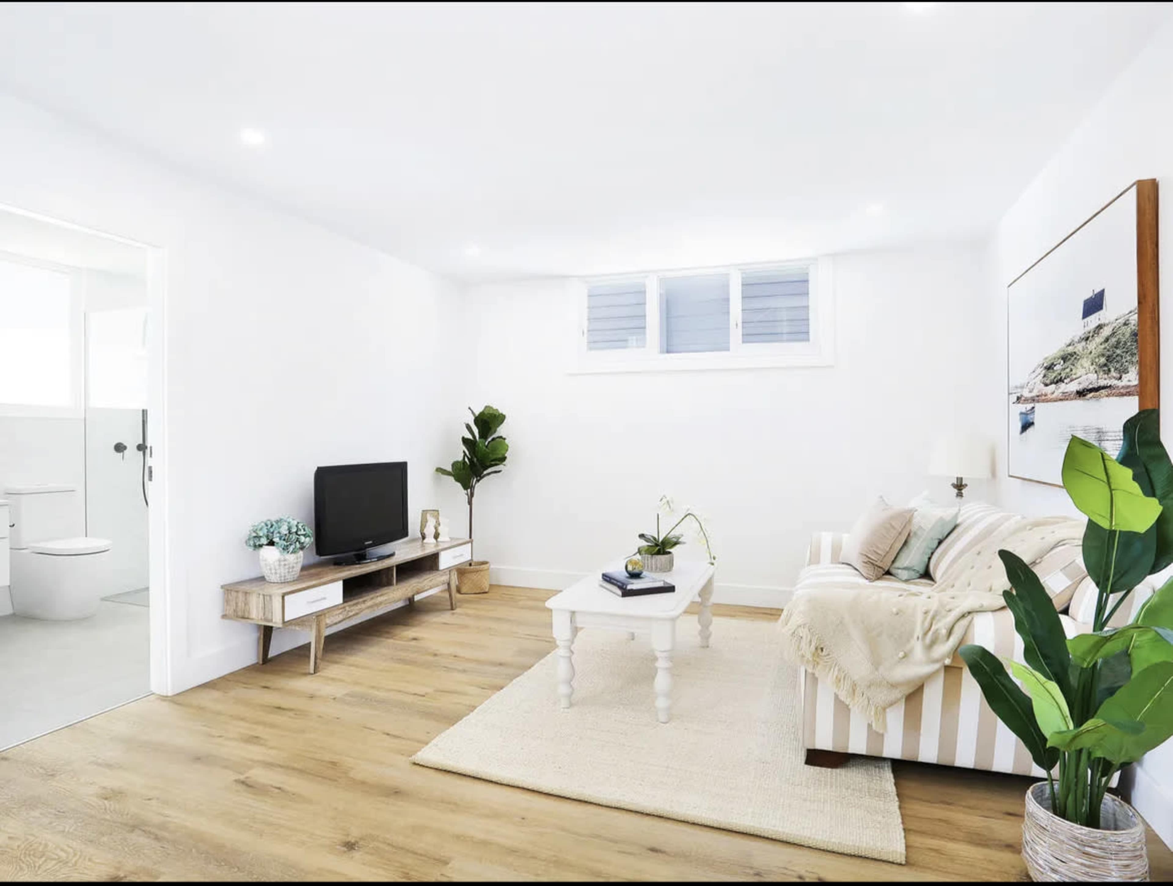 A bright living room features a white couch, a wooden coffee table, a television on a stand, and a potted plant, with a bathroom visible through an open door.