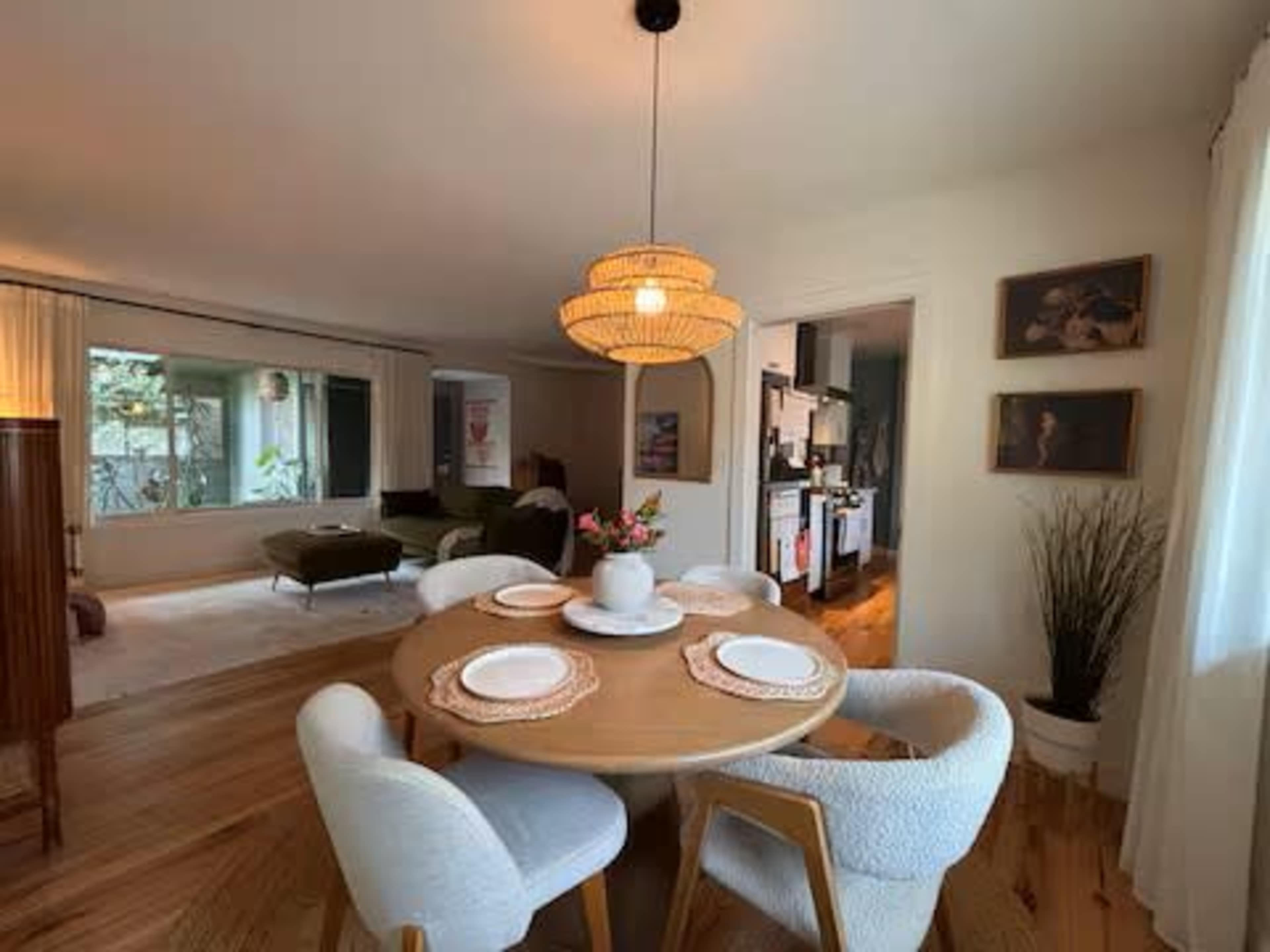 A round dining table with white plates sits at the center of a well-lit room featuring a window, a kitchen, and decorative artwork on the walls.