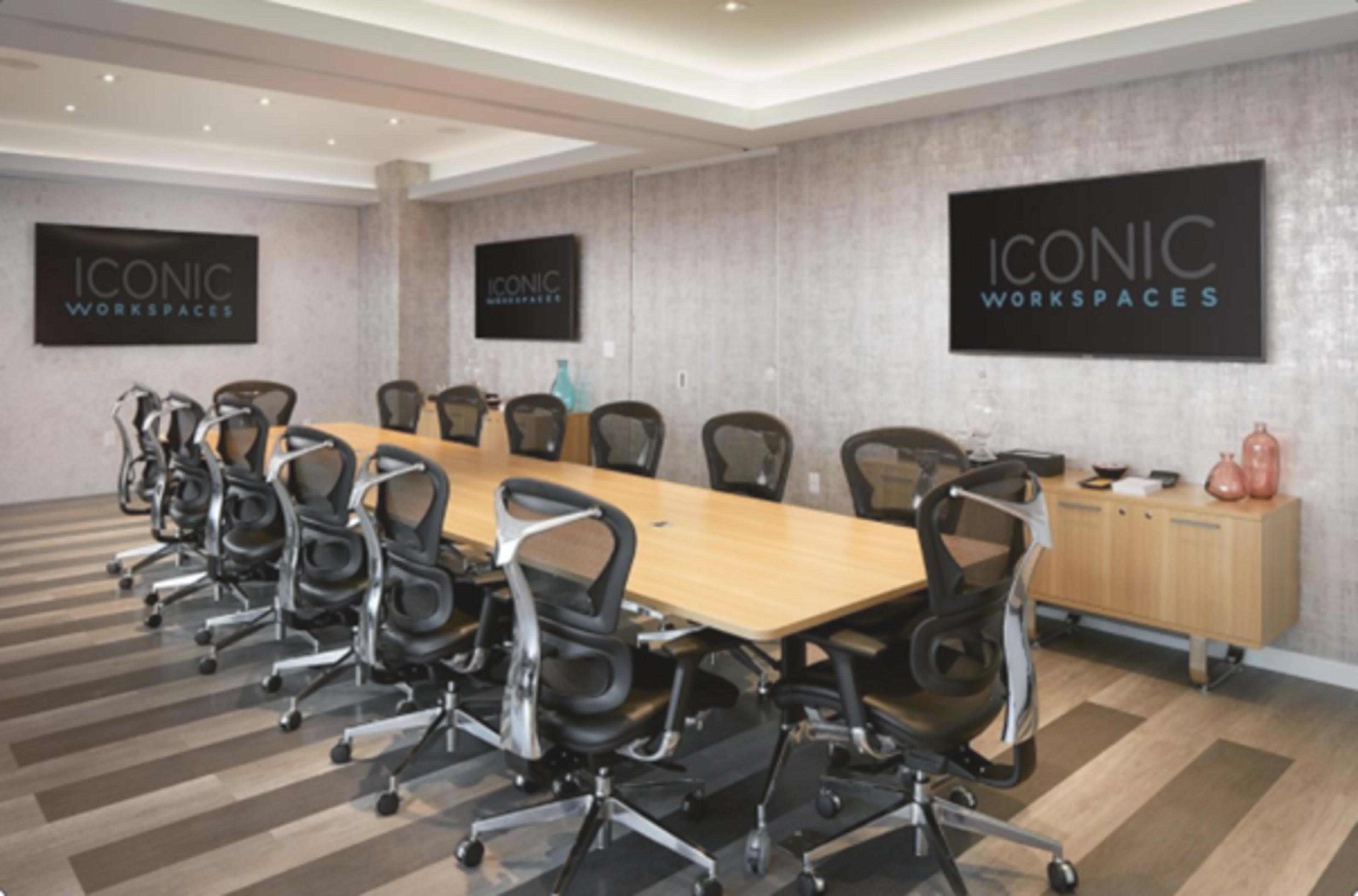 A modern conference room features a long wooden table surrounded by ergonomic black chairs, with large screens displaying the text "ICONIC WORKSPACES" on the walls.