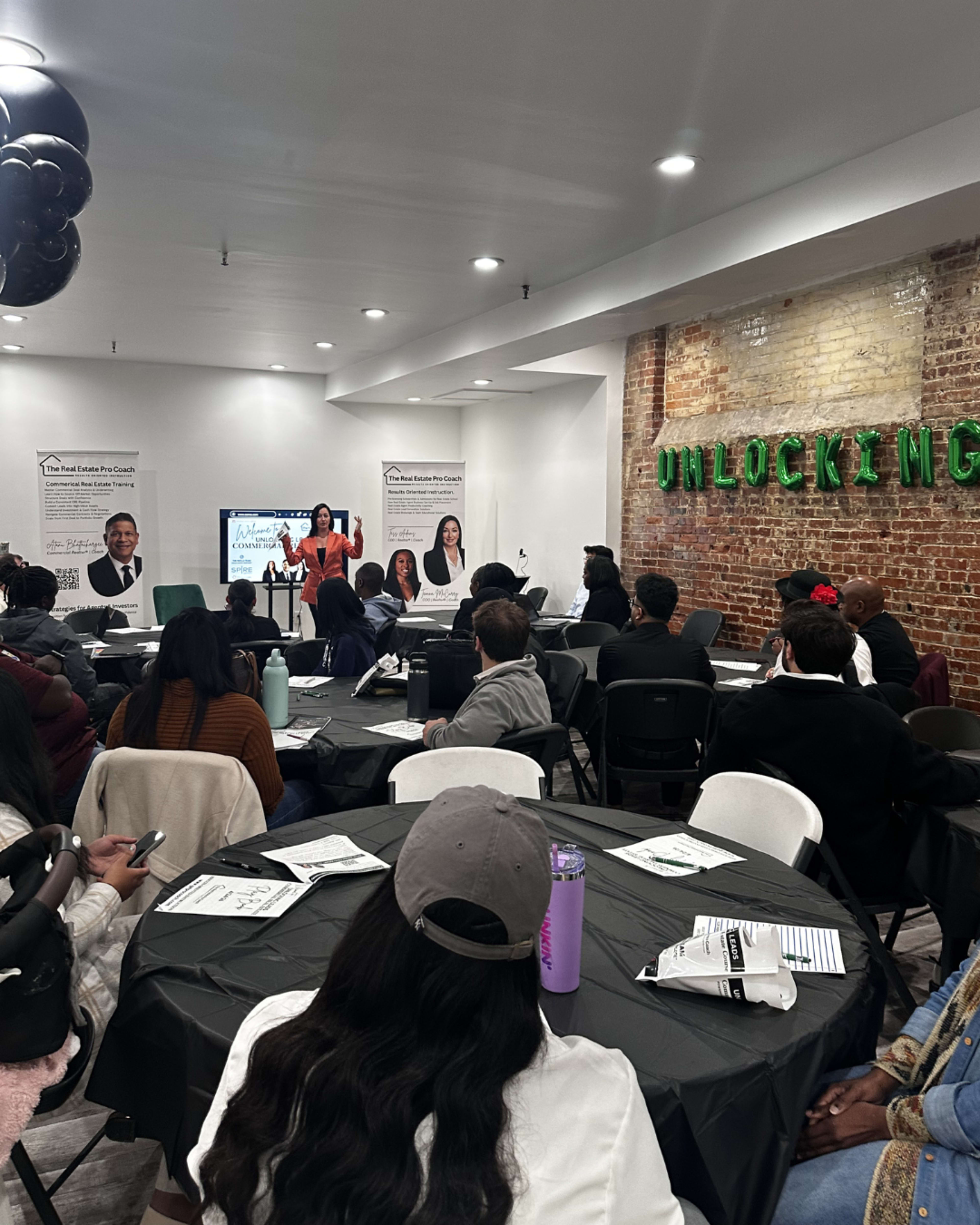 A group of people is seated at round tables in a room with brick walls, listening to a speaker presenting on a screen.
