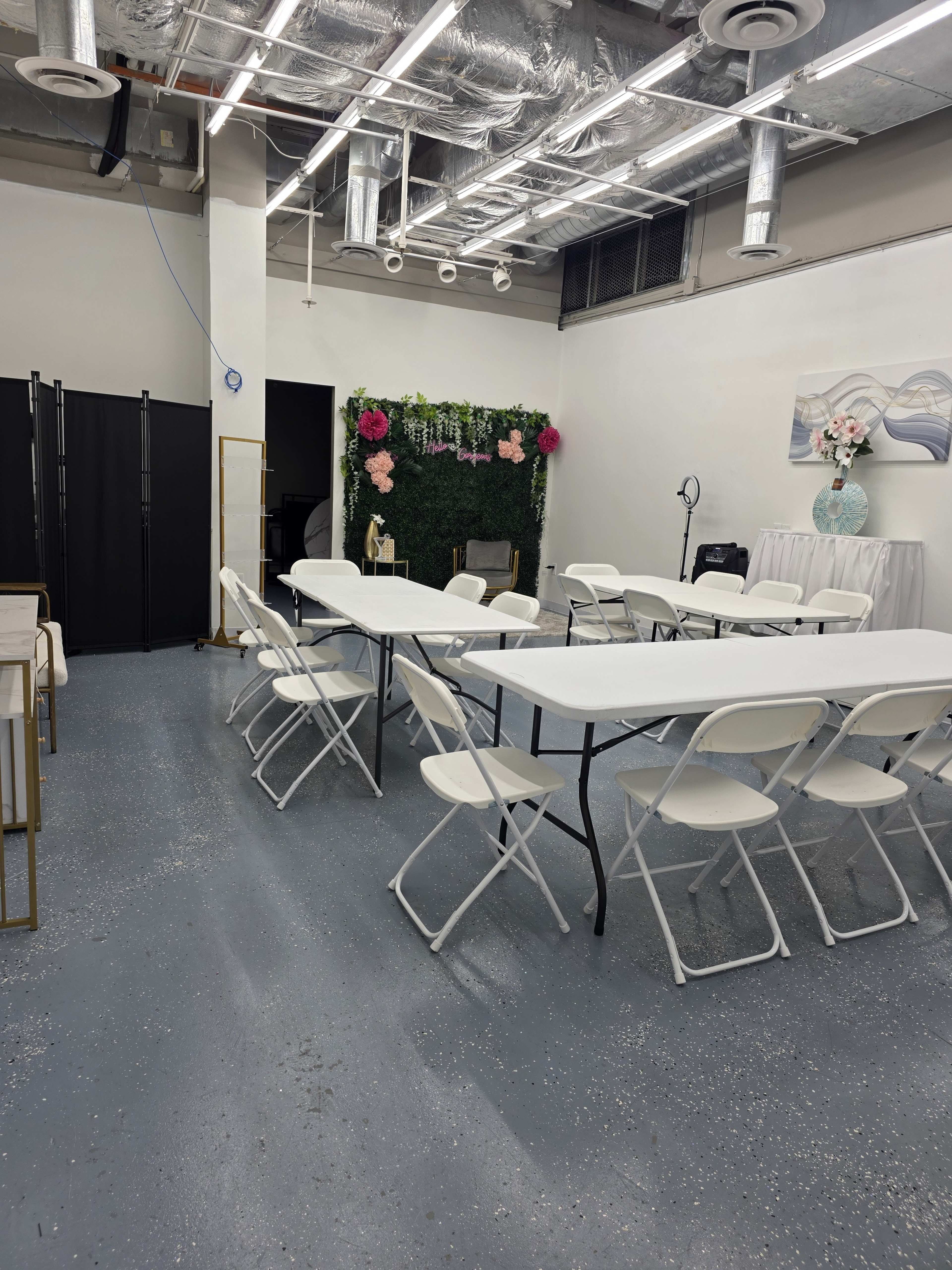 A spacious room with white folding tables and chairs arranged for an event, featuring greenery and decorations on one wall.