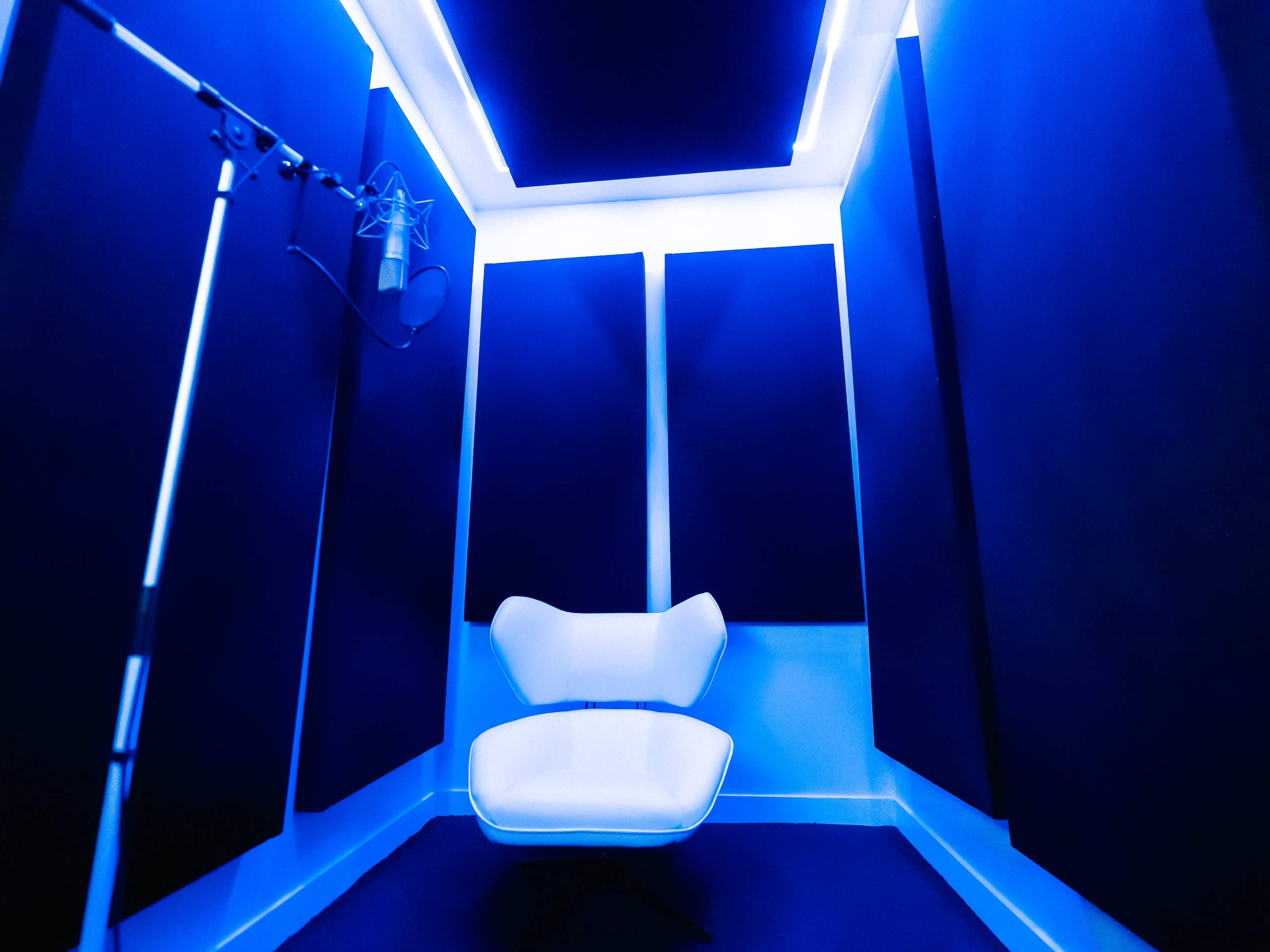A soundproof recording studio featuring a white chair and a microphone on a stand, illuminated by blue lighting.