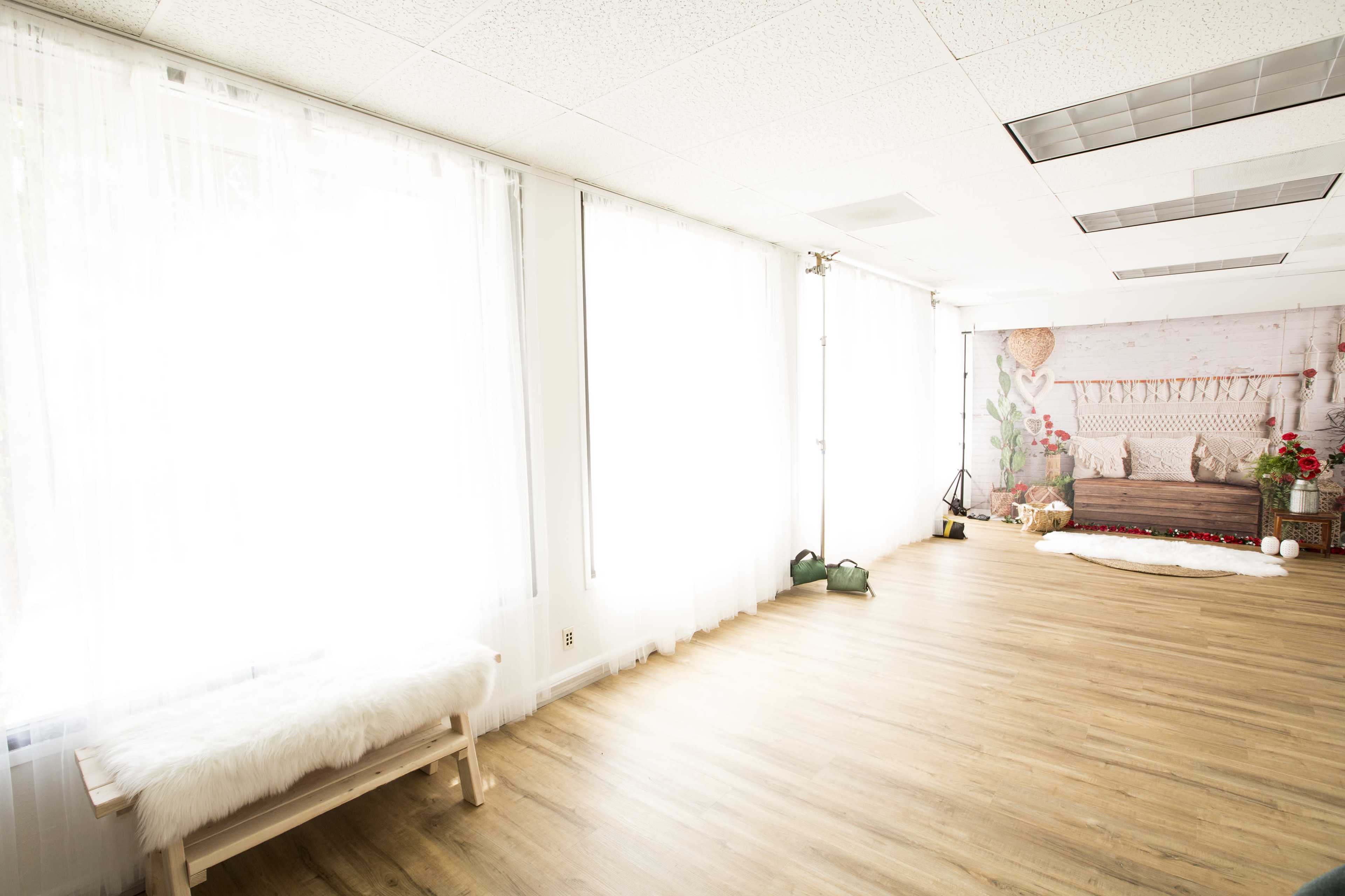 The image shows a spacious room with large windows covered by sheer curtains, a wooden bench, and a decorative area featuring a bed with floral accents.