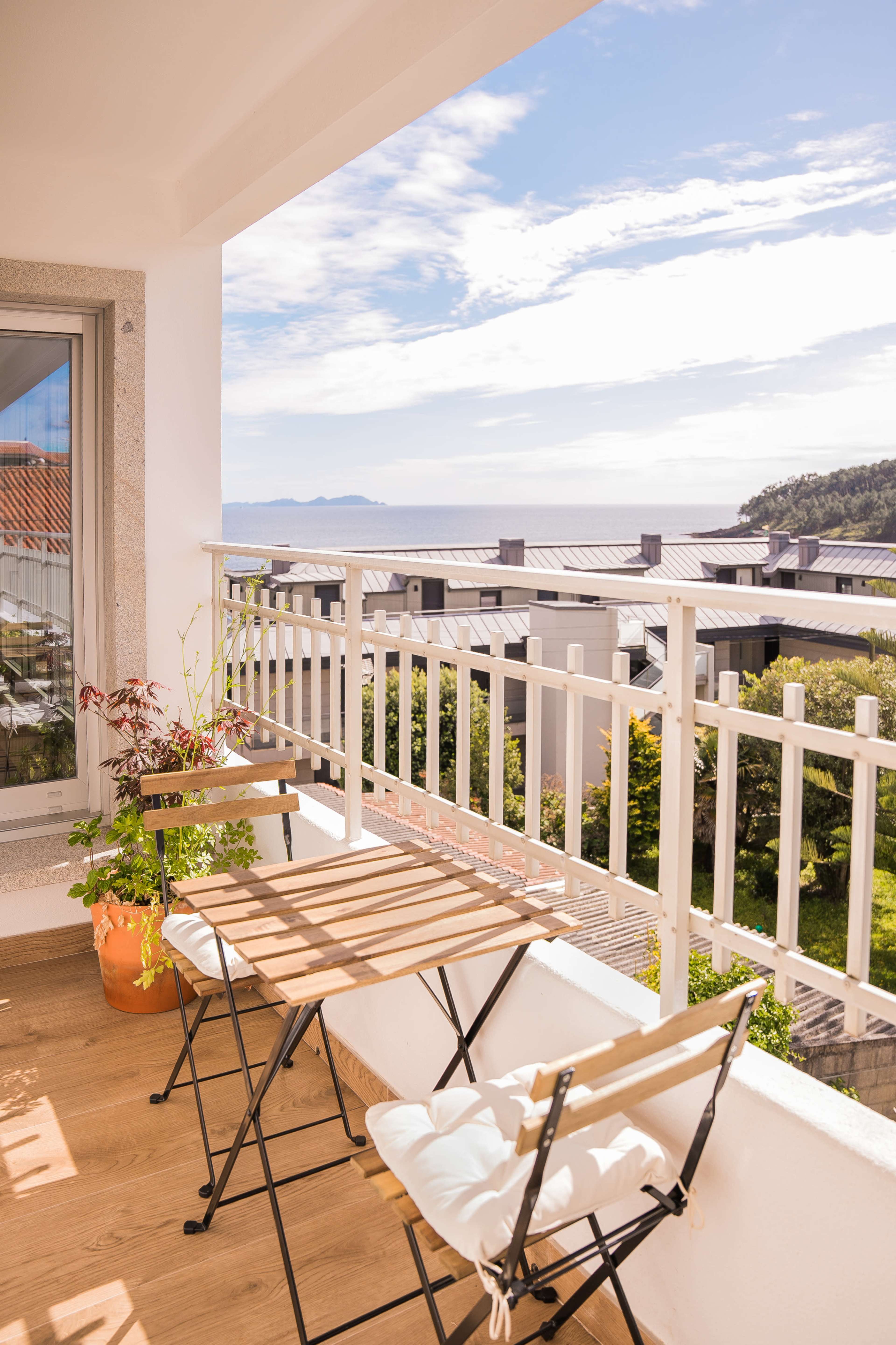 A wooden balcony with two folding chairs overlooks a coastal view featuring water and distant hills.