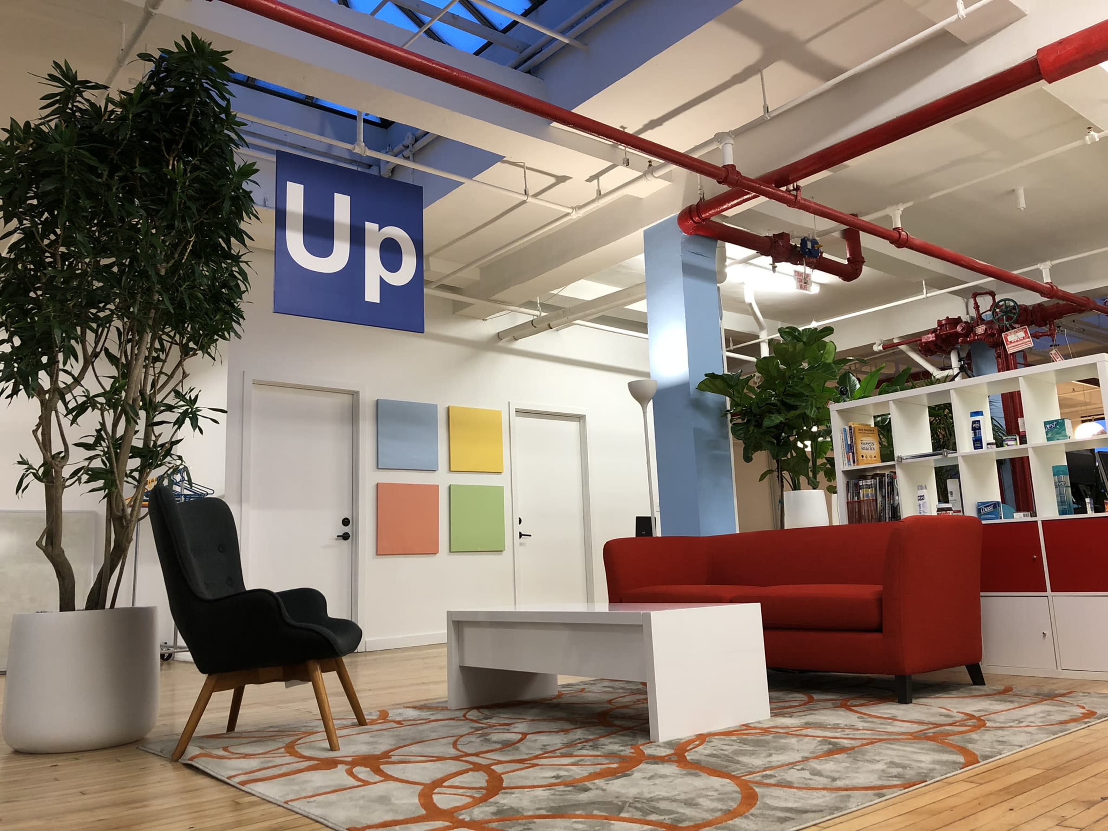 A modern office space with a red sofa, a black chair, a white coffee table, colorful wall panels, and greenery.