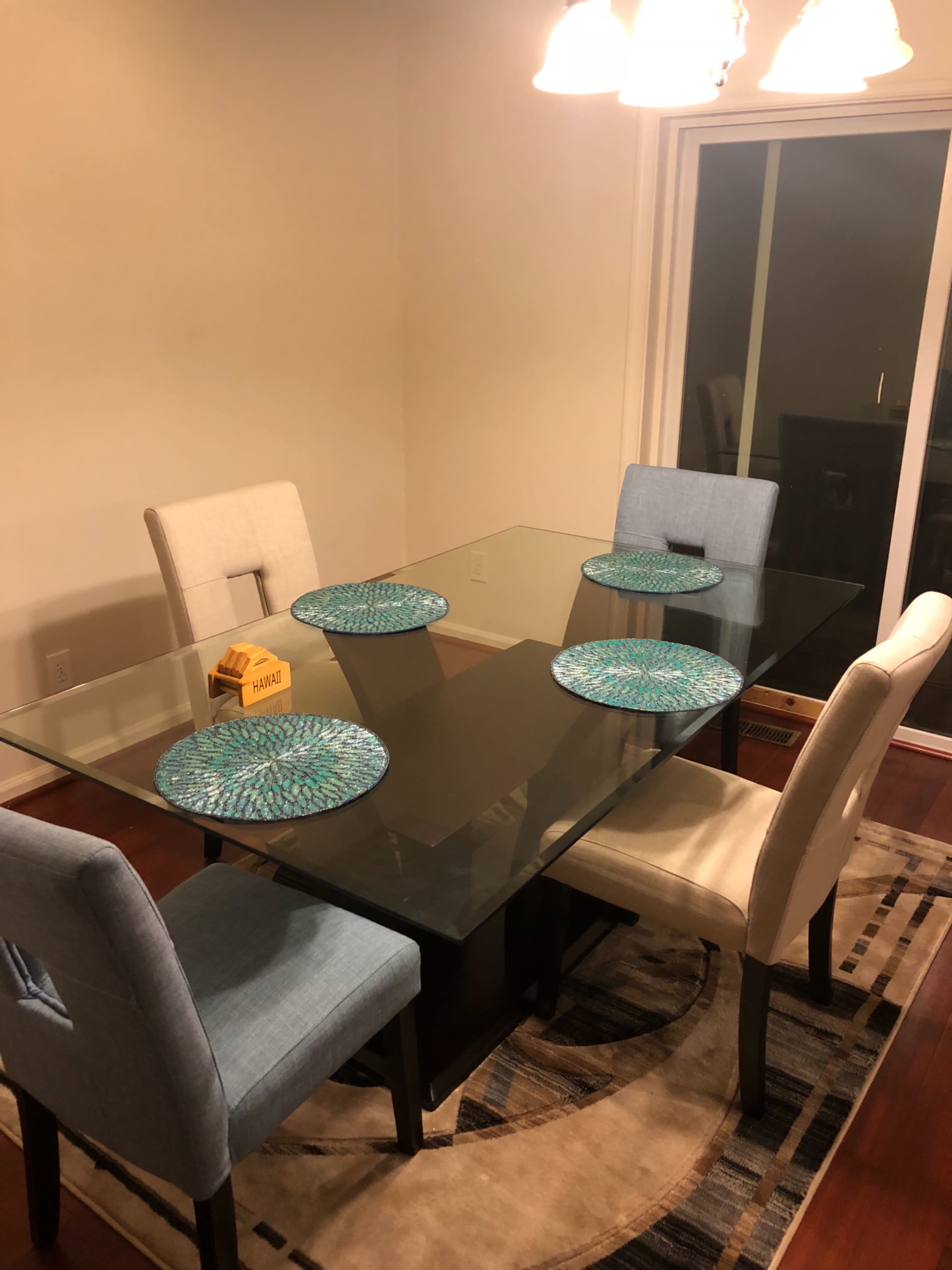 A rectangular glass dining table is set for four with round placemats, surrounded by four chairs in a well-lit room.