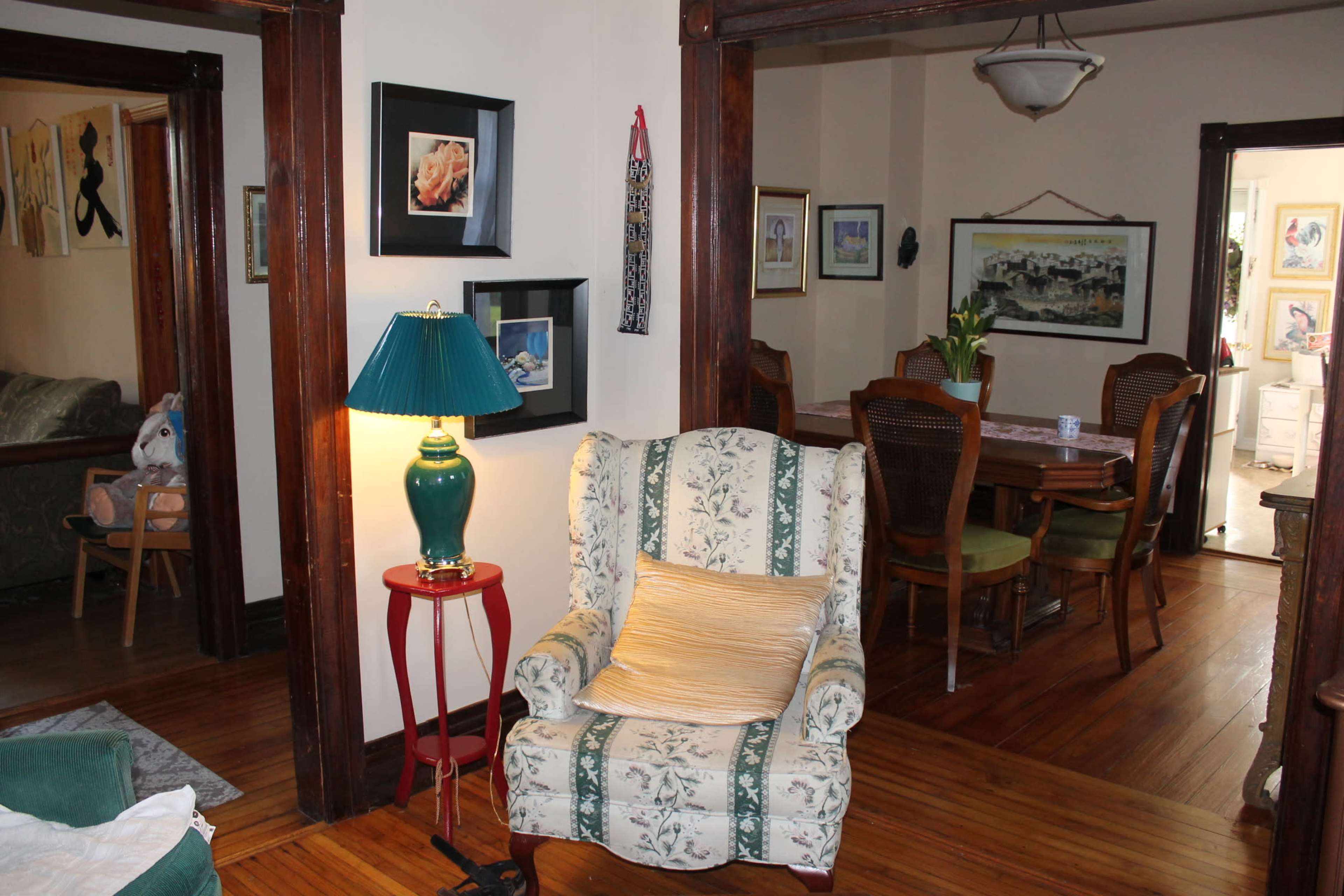 A patterned armchair sits next to a table lamp in a room with wooden trim, while a dining area is visible in the background.