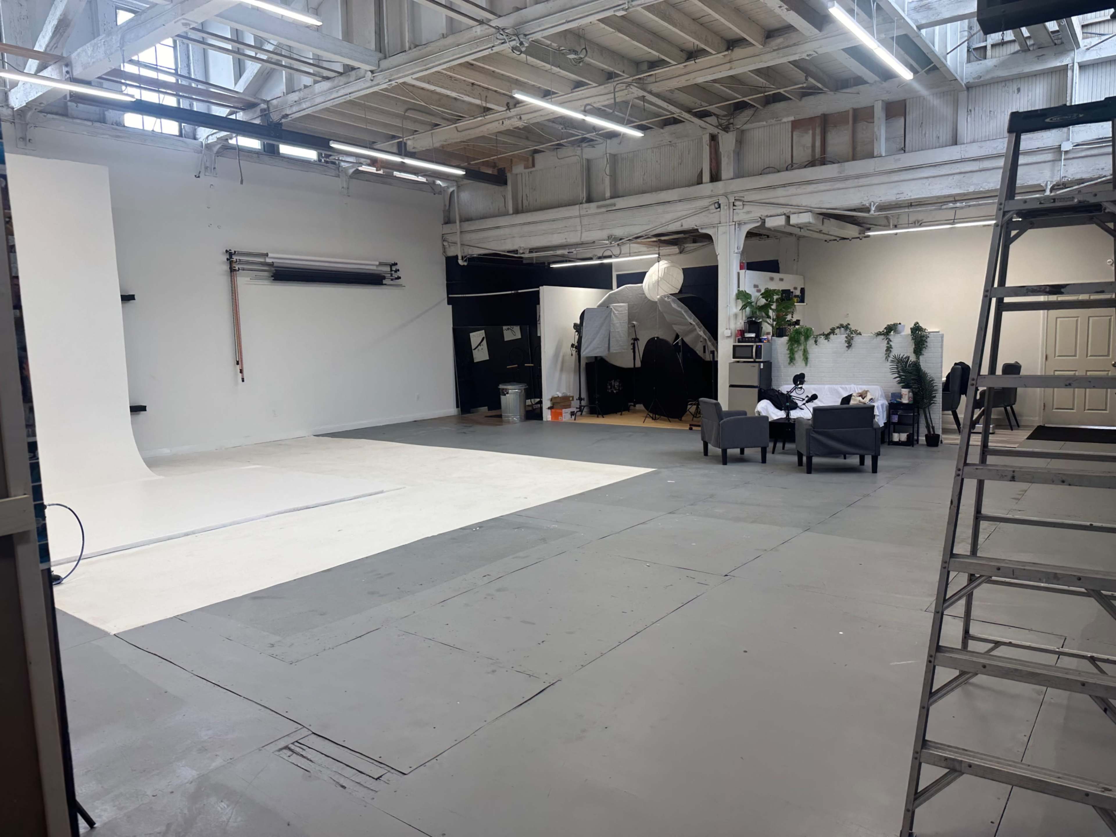 The image shows a spacious photography studio with a white backdrop, a seating area, and various equipment scattered around.