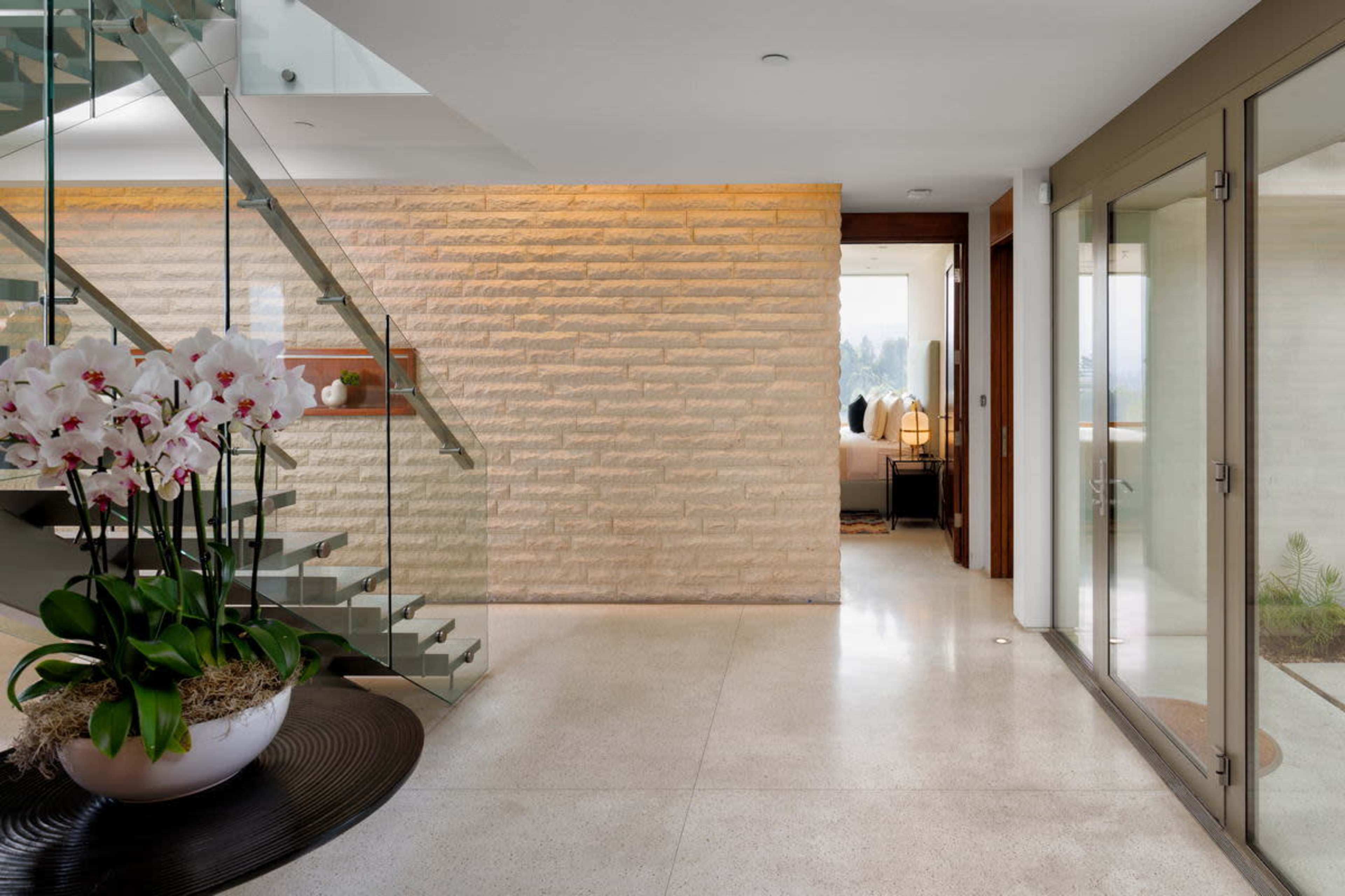 The image depicts a modern interior entrance featuring a staircase, a large planter with white orchids, and a glass door leading outside.