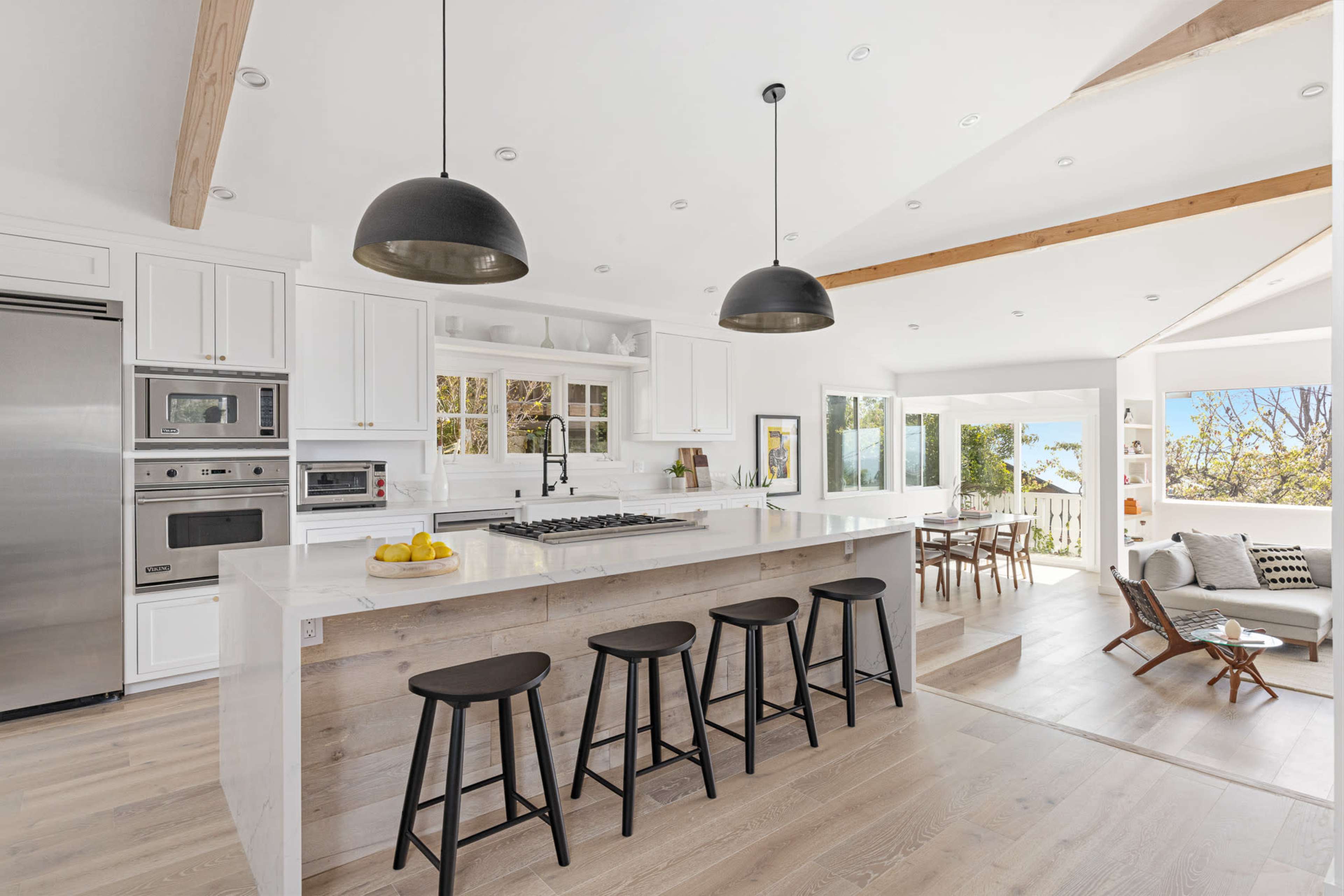 A modern kitchen features white cabinetry, a large island with seating, and large windows that provide a view of the outdoors.