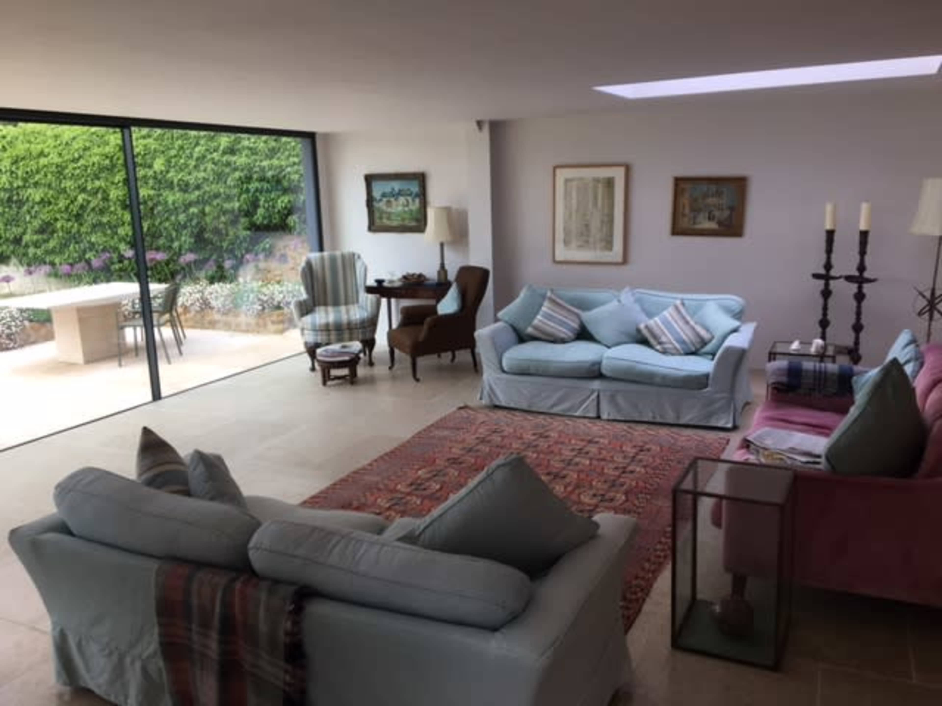 The spacious living room features light-colored sofas, a patterned rug, and large windows that provide a view of the garden outside.