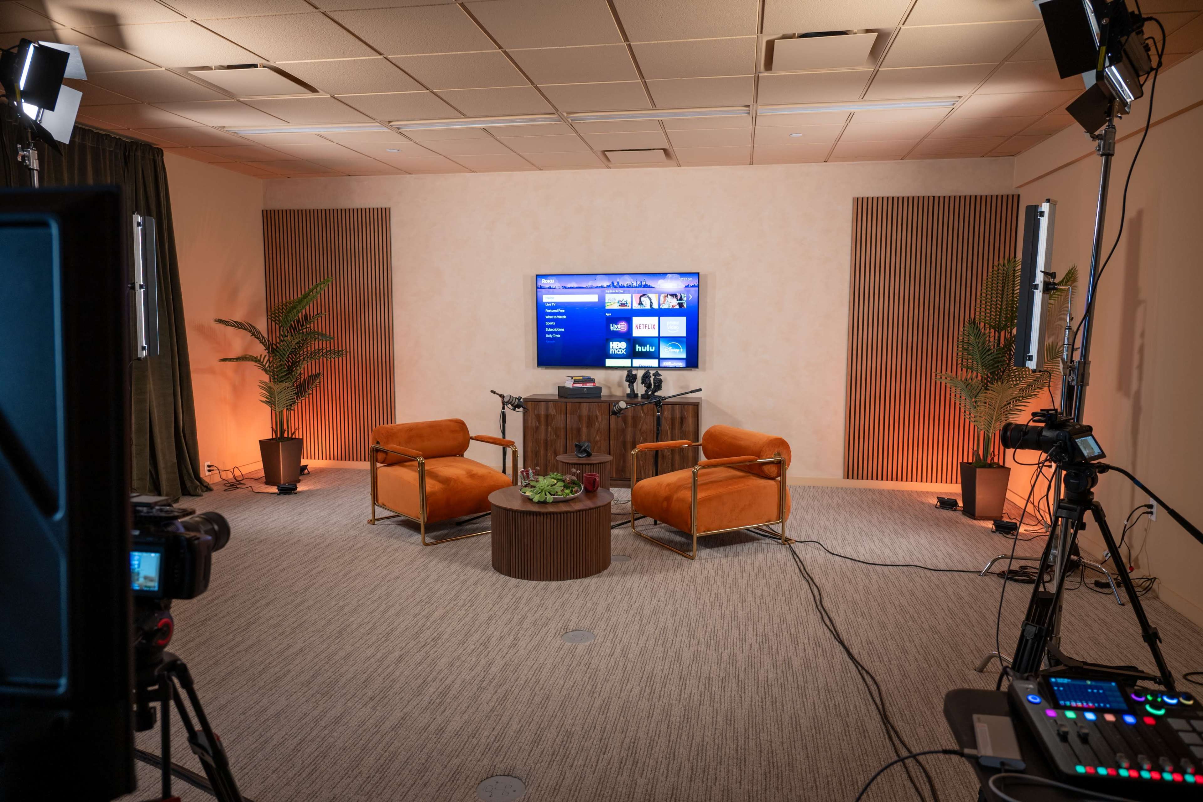 The image shows a studio setup with two orange chairs facing a coffee table, a television on the wall, and camera equipment positioned around the room.