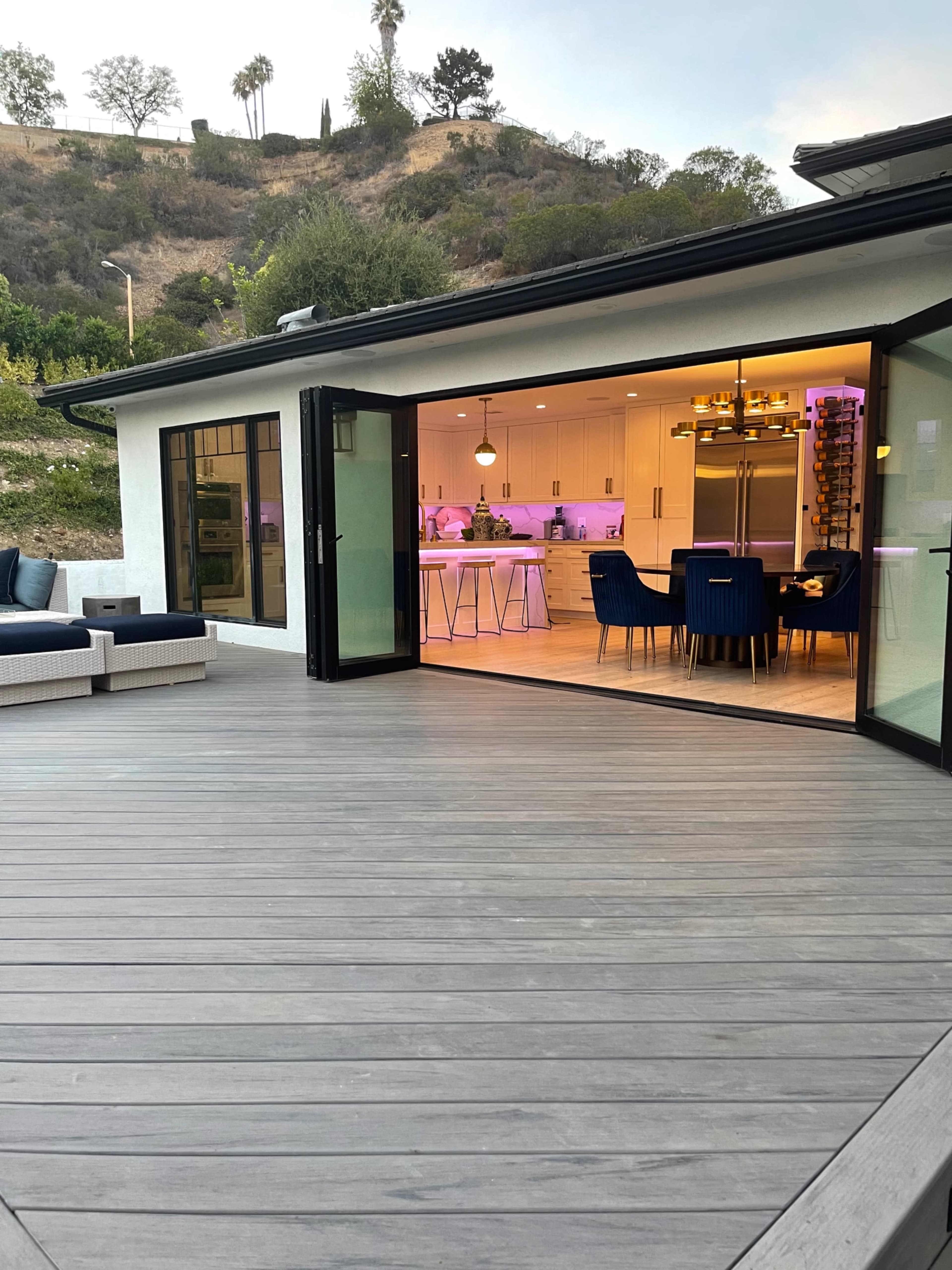 A modern home with large glass doors leading to a deck, overlooking a hillside with trees, and an interior kitchen area illuminated with colorful lighting.