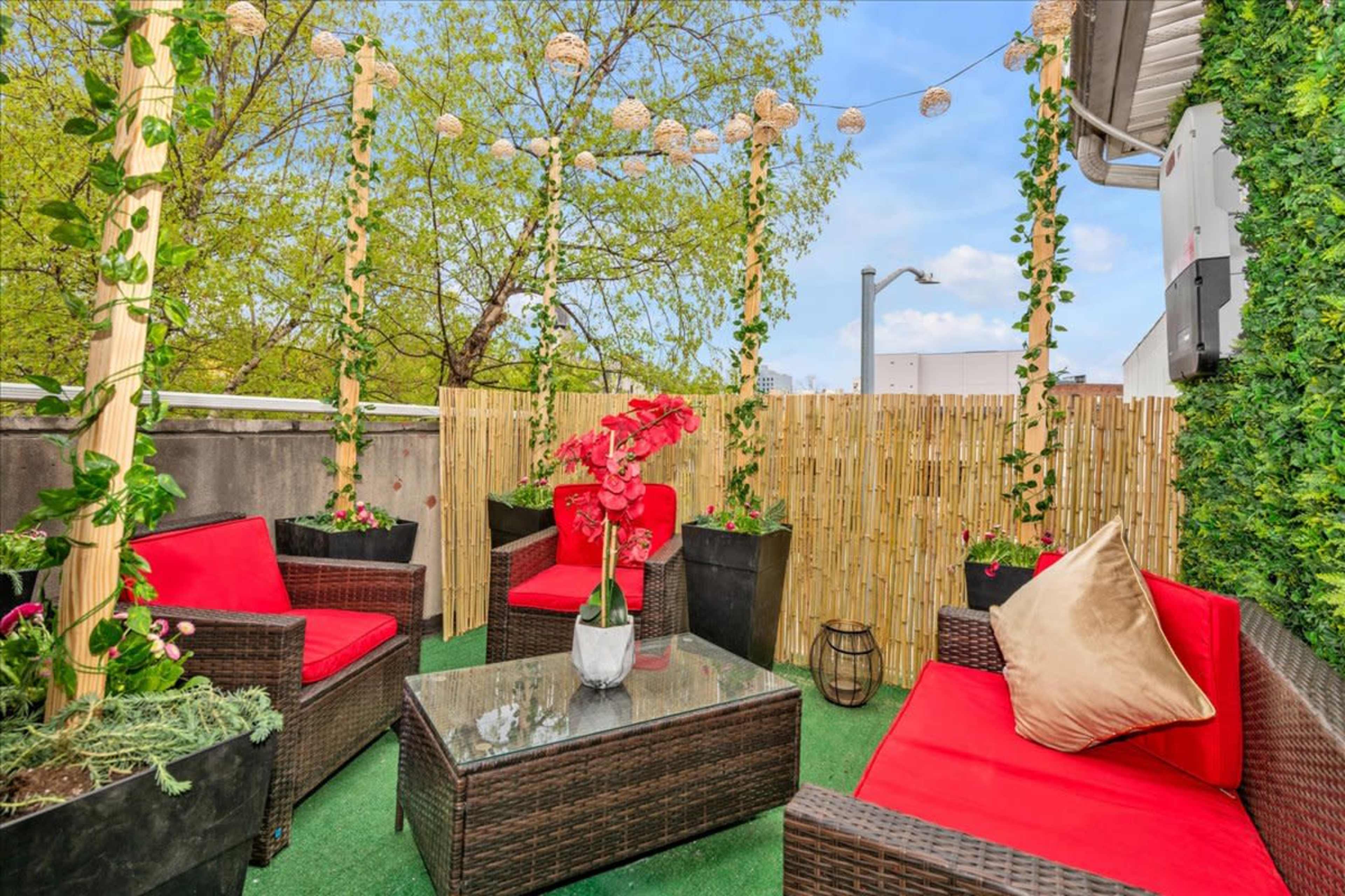 A rooftop terrace with red outdoor furniture, potted plants, and decorative lighting against a backdrop of greenery.
