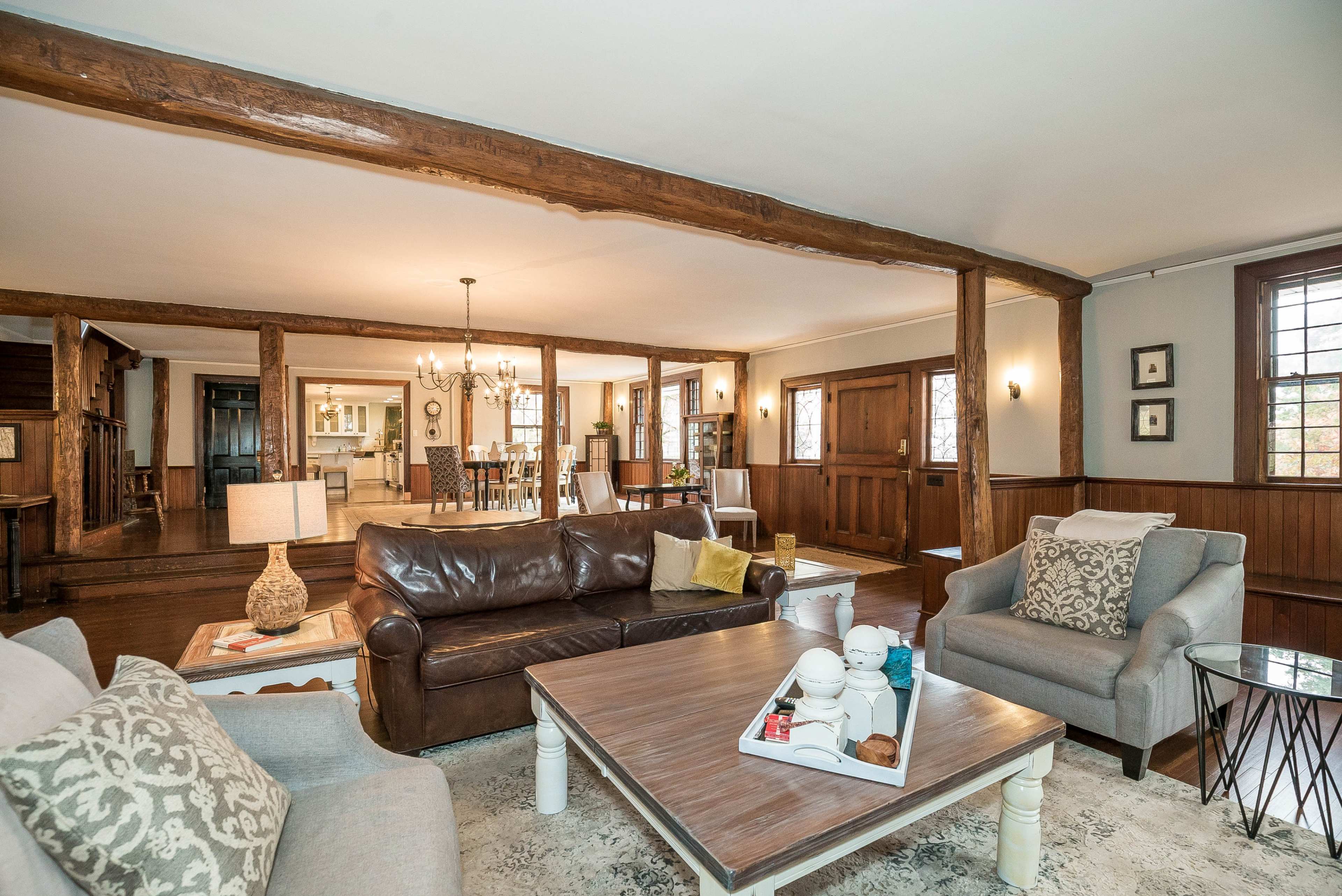 A spacious living room with wooden beams, a mix of seating arrangements, and a dining area visible in the background.