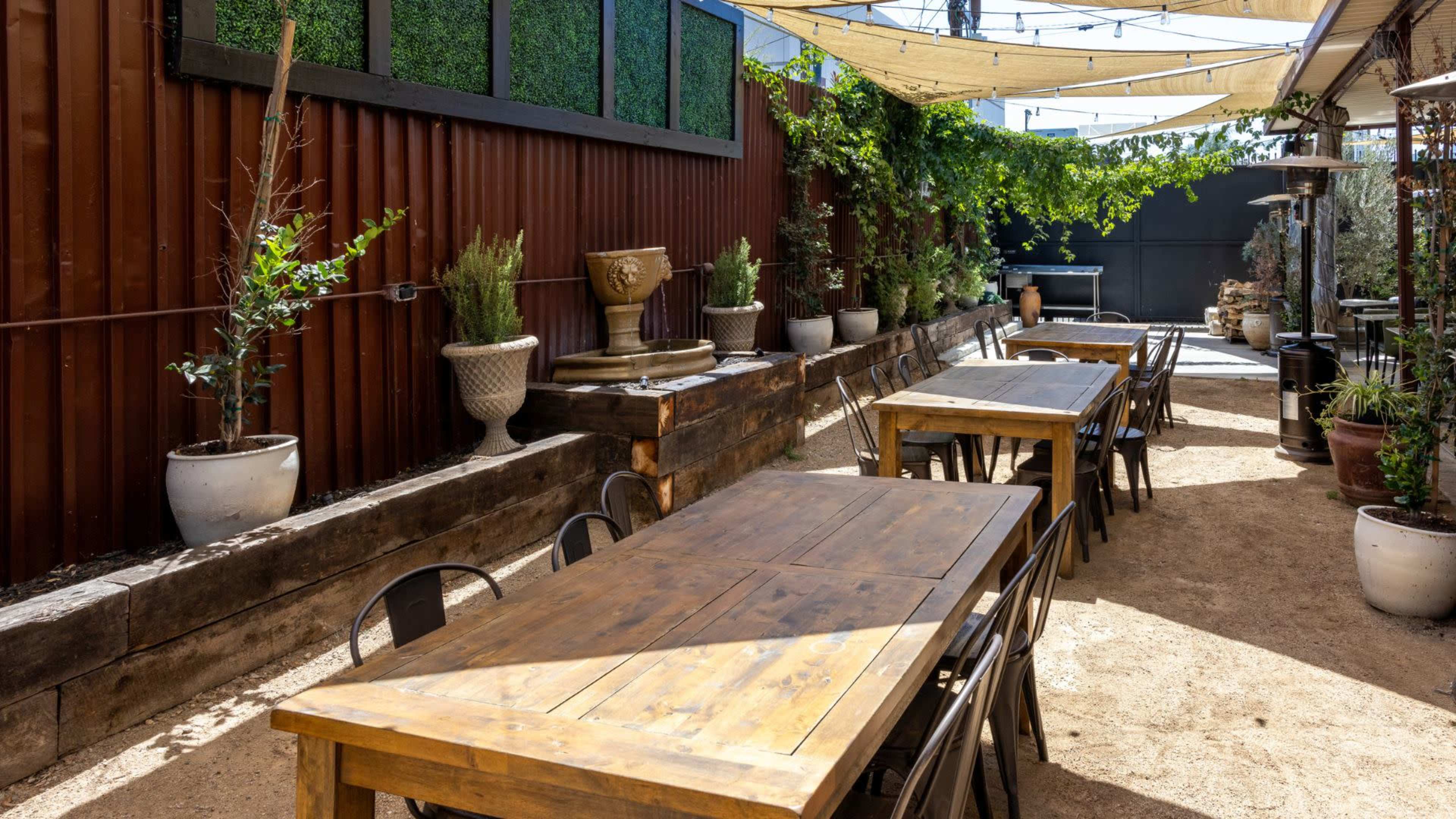 A narrow outdoor patio features wooden tables, chairs, and planters with greenery, surrounded by rustic fencing and a wall adorned with plants.
