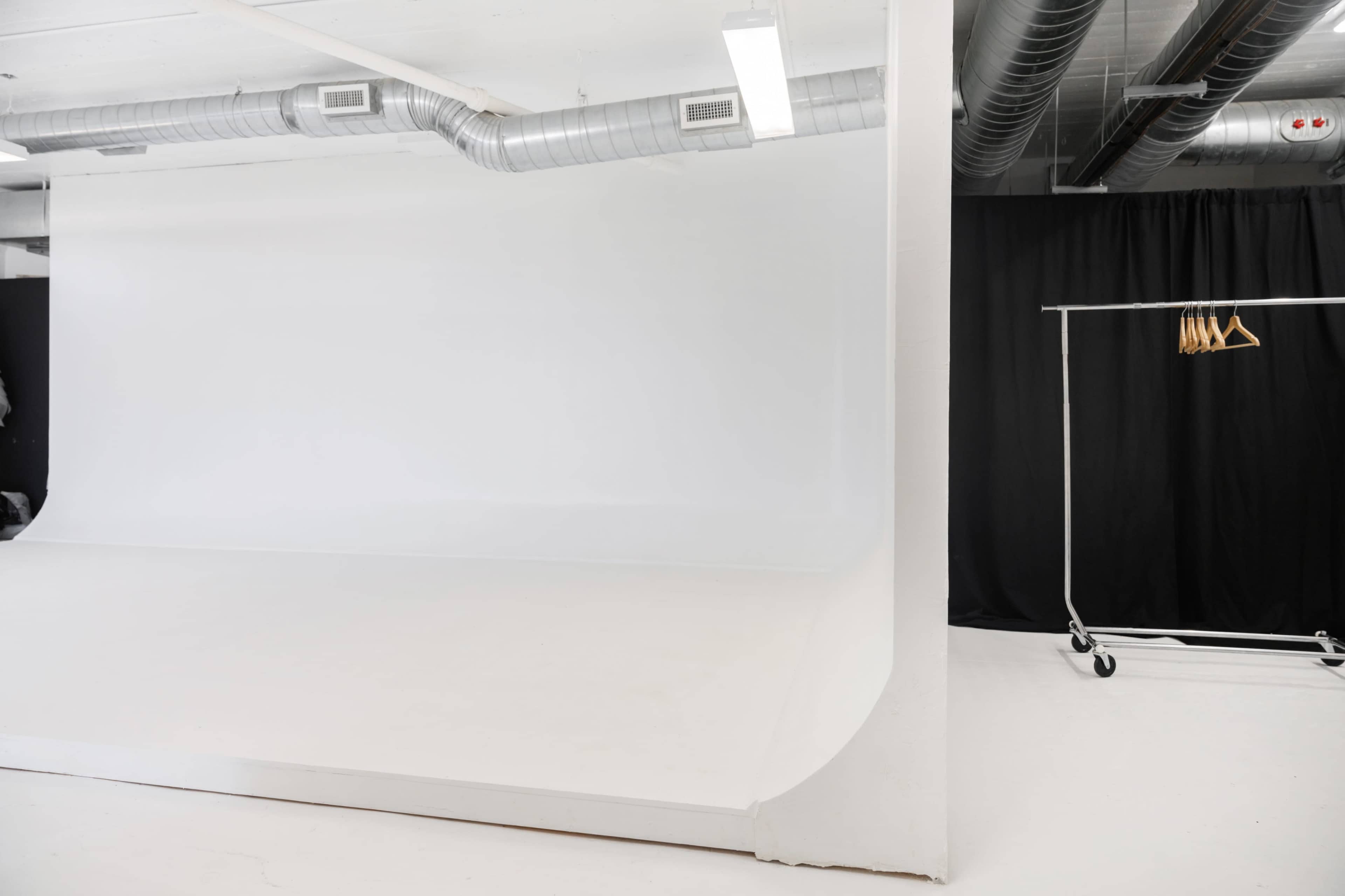 A photography studio with a large white backdrop and a clothing rack on wheels with hangers against a black curtain.