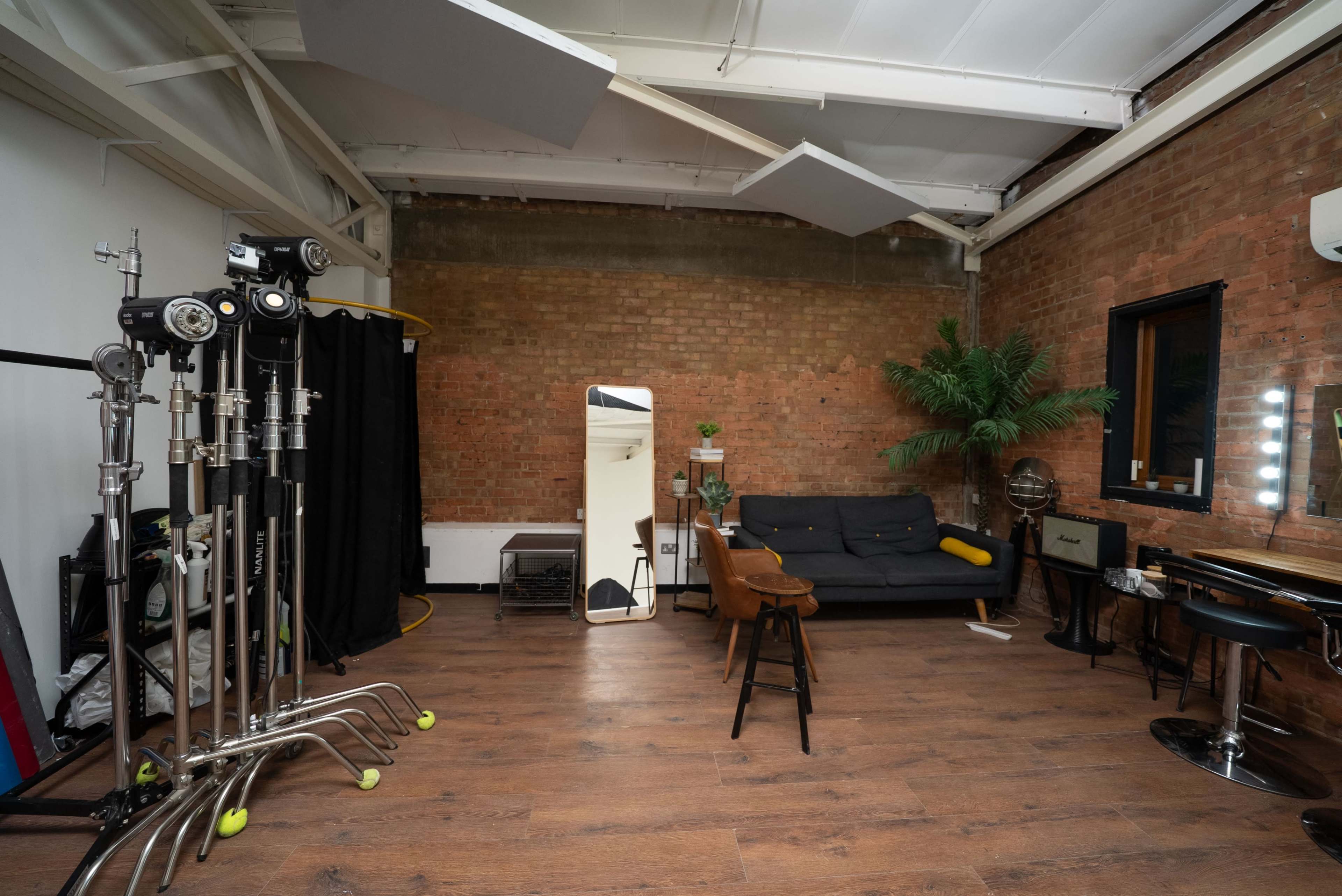 The image shows a spacious studio with exposed brick walls, a dark sofa, a mirror, and various photography equipment arranged around the room.