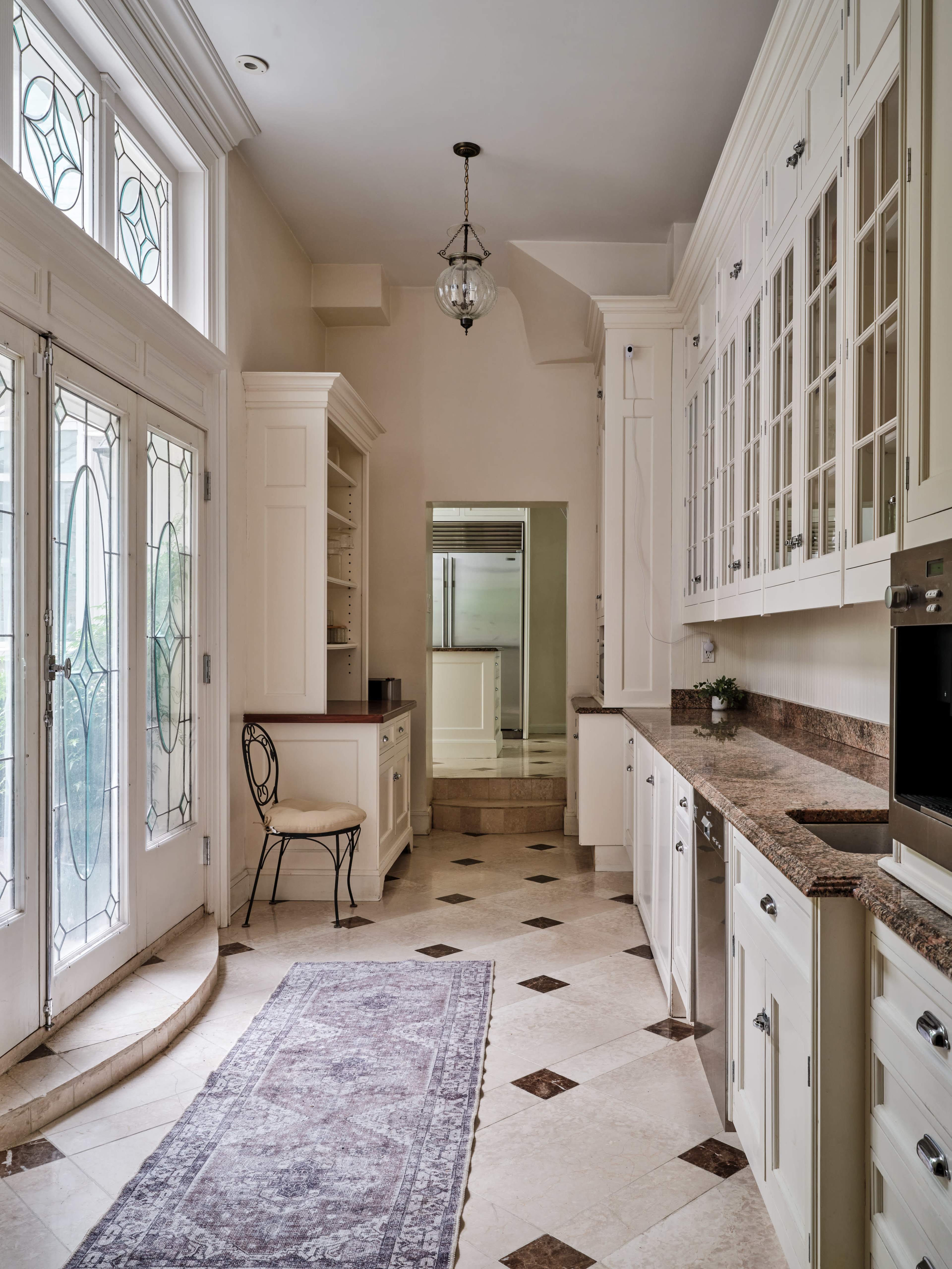 A spacious kitchen with white cabinetry, a stone countertop, and a patterned rug on the tiled floor, leading to large glass doors and a mirror in the background.