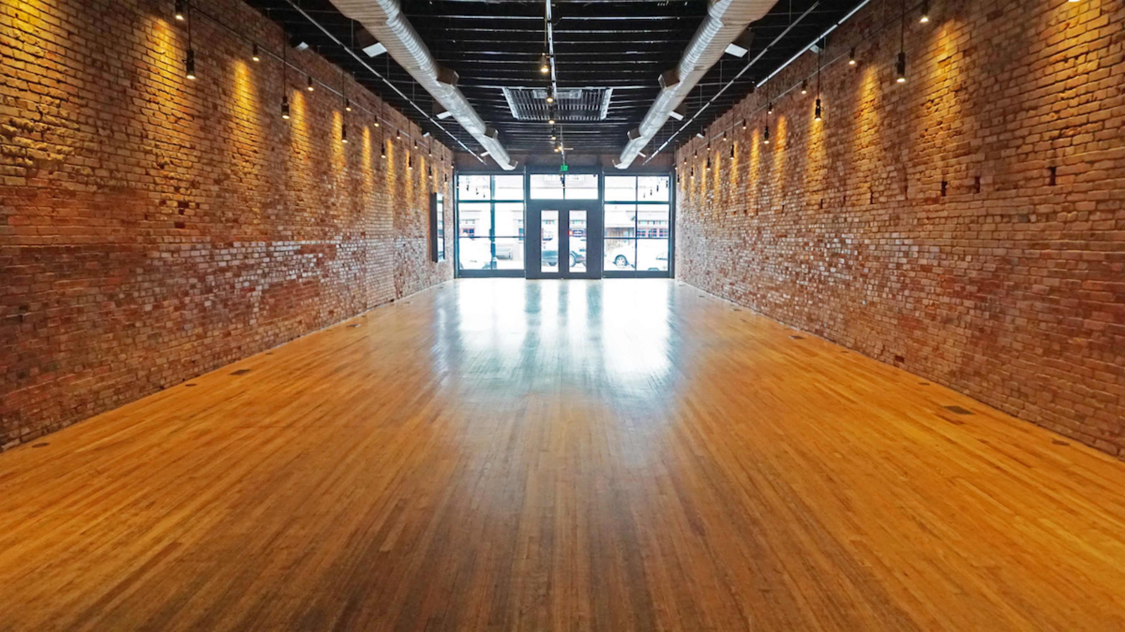 A long, empty room with exposed brick walls and polished wooden floors, illuminated by overhead lights.