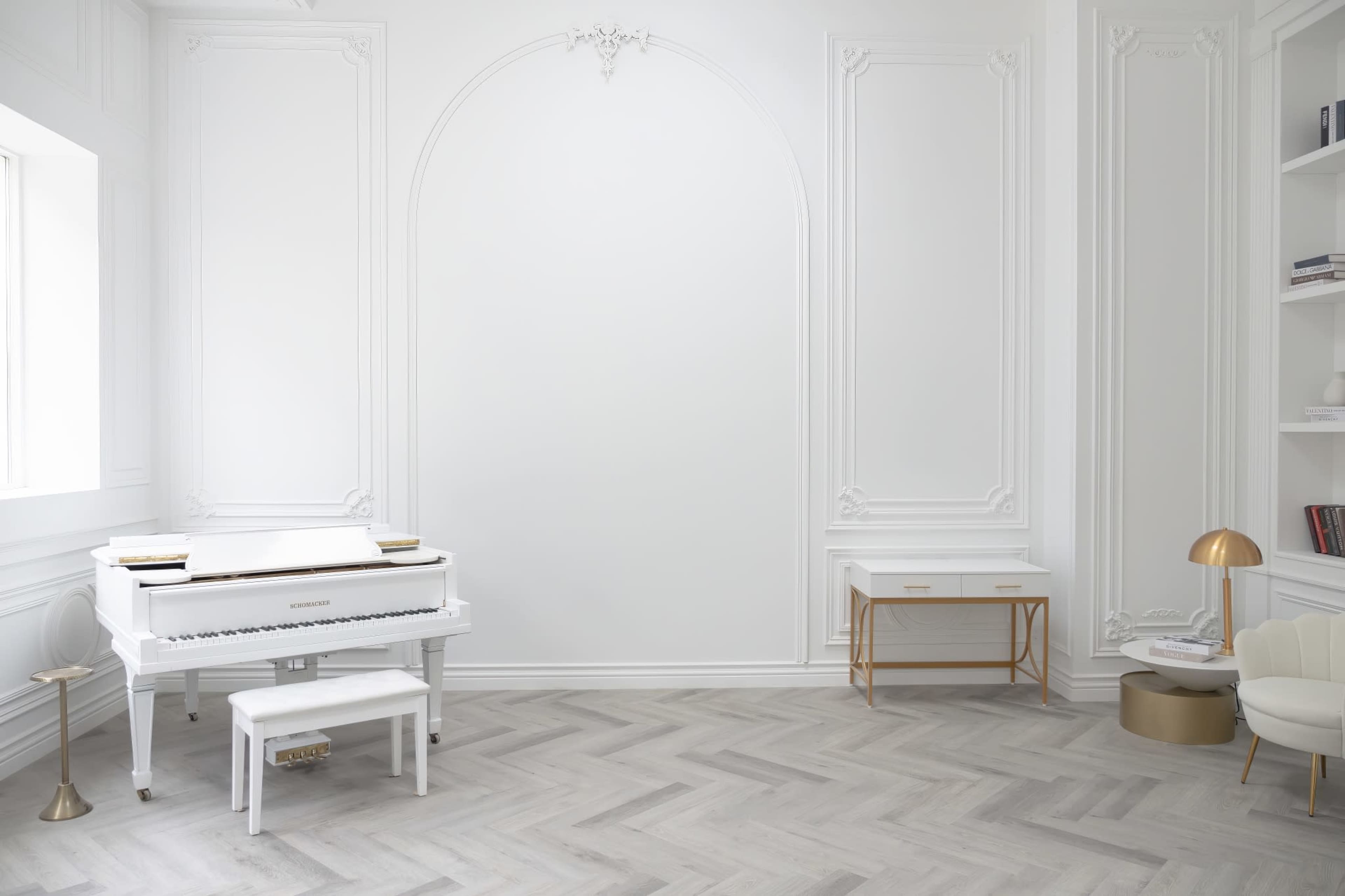 The room features a white grand piano, a modern desk, and a cream-colored chair against a light-colored, intricately designed wall.