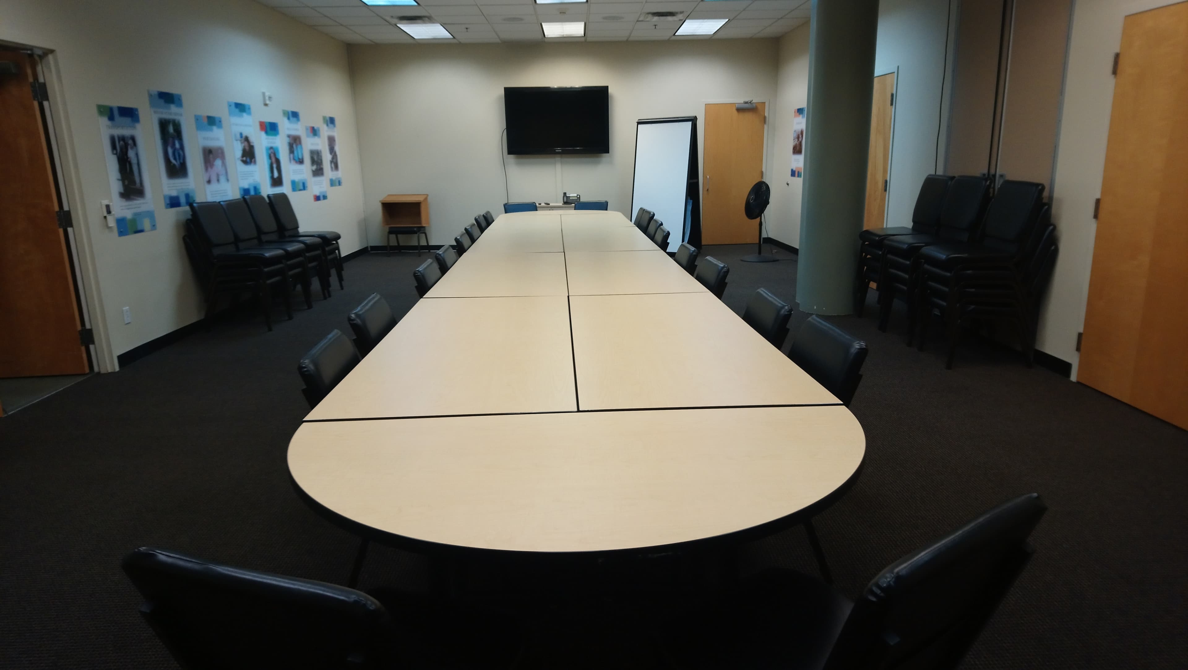 A conference room featuring a large, rectangular table surrounded by black chairs, with a television and whiteboard on one wall.
