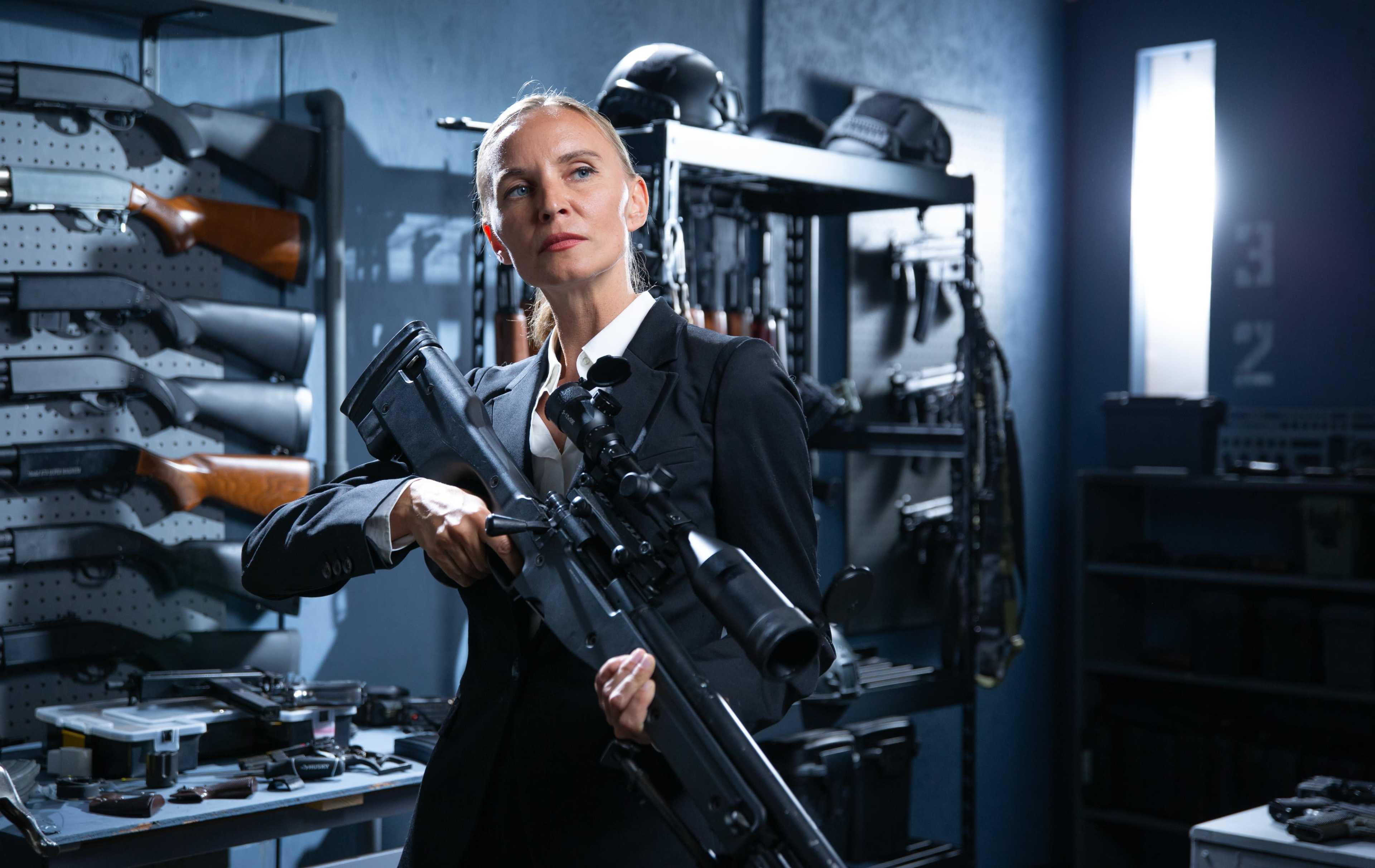 A woman in formal attire holds a rifle while standing in a room filled with various firearms and tactical equipment.