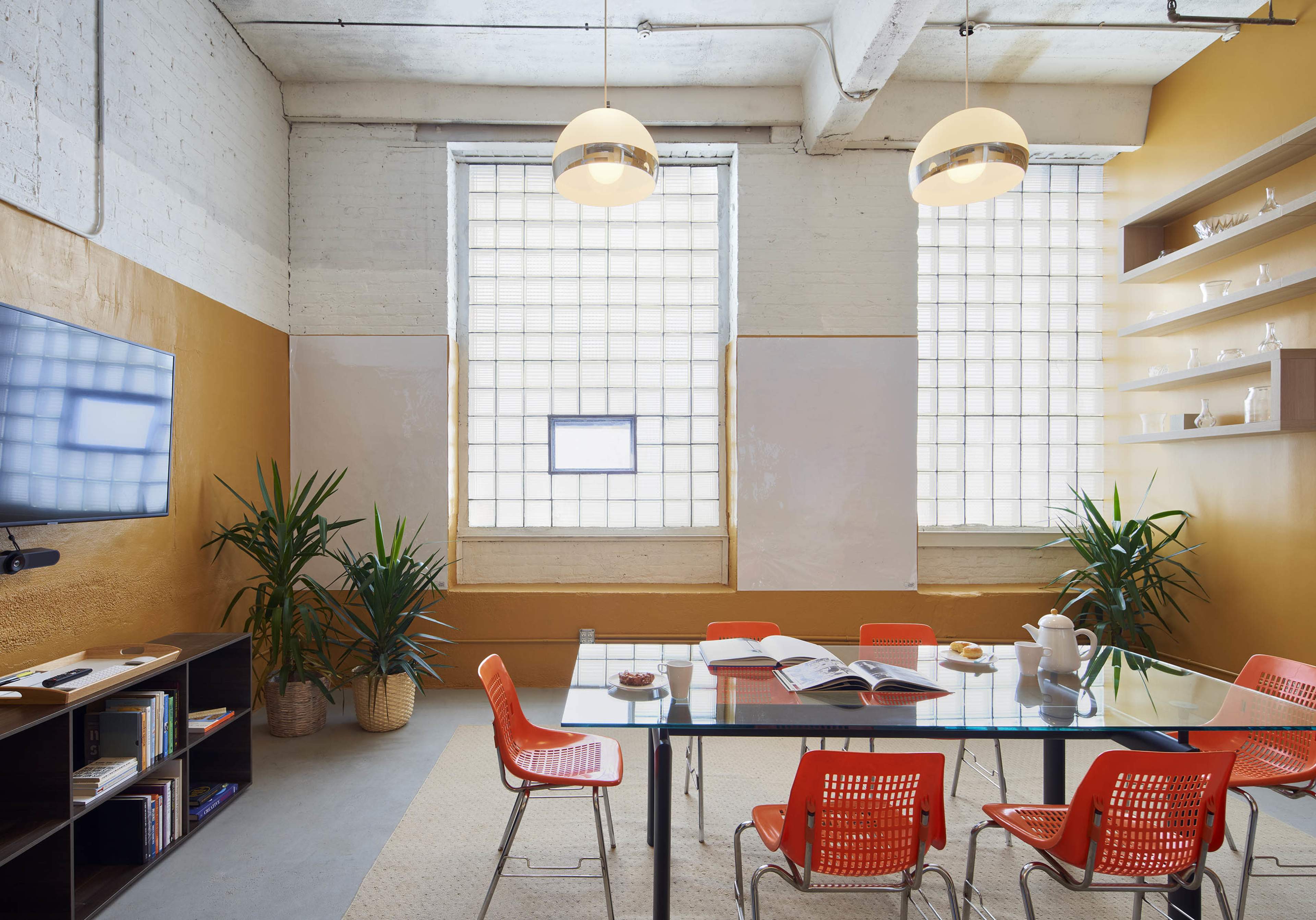 The image shows a modern meeting room with a glass table surrounded by orange chairs, two large windows, and a cabinet with books and decorative items.