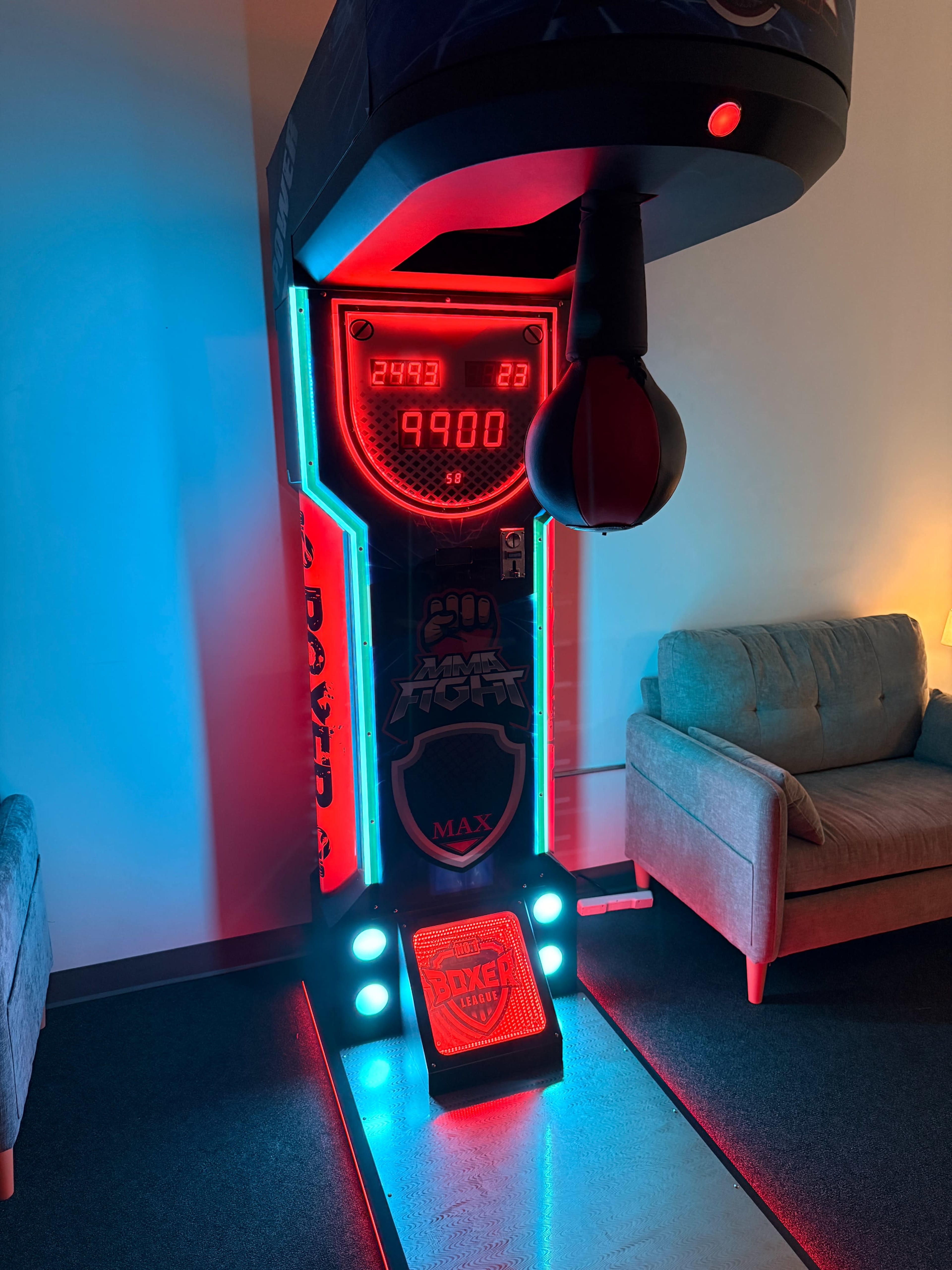 The image shows a brightly lit punching bag arcade game with a digital score display and a striking red and blue color scheme, placed in a room with two light-colored sofas.