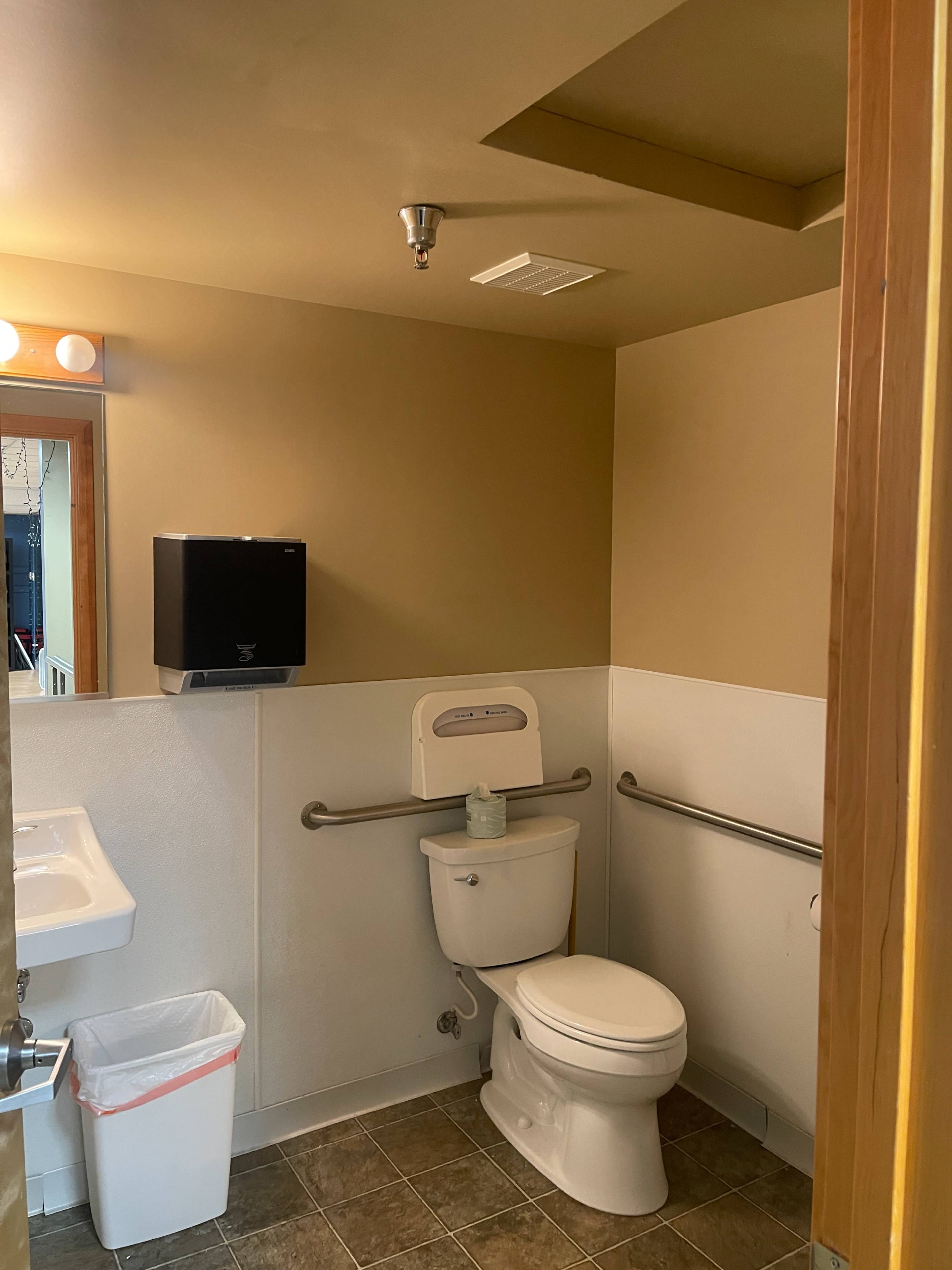 The image shows a small bathroom with a toilet, a sink, a hand dryer, and a trash can.