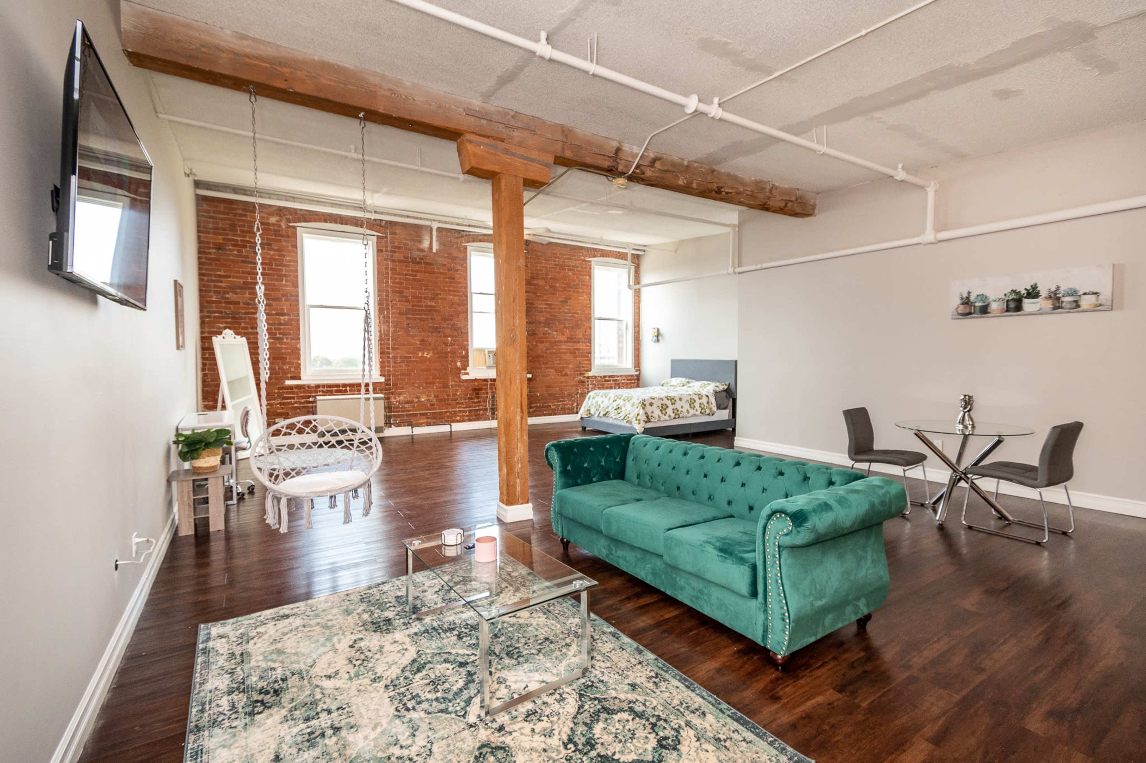 A spacious, modern living area with a green velvet sofa, a glass table with chairs, and exposed brick walls featuring large windows.