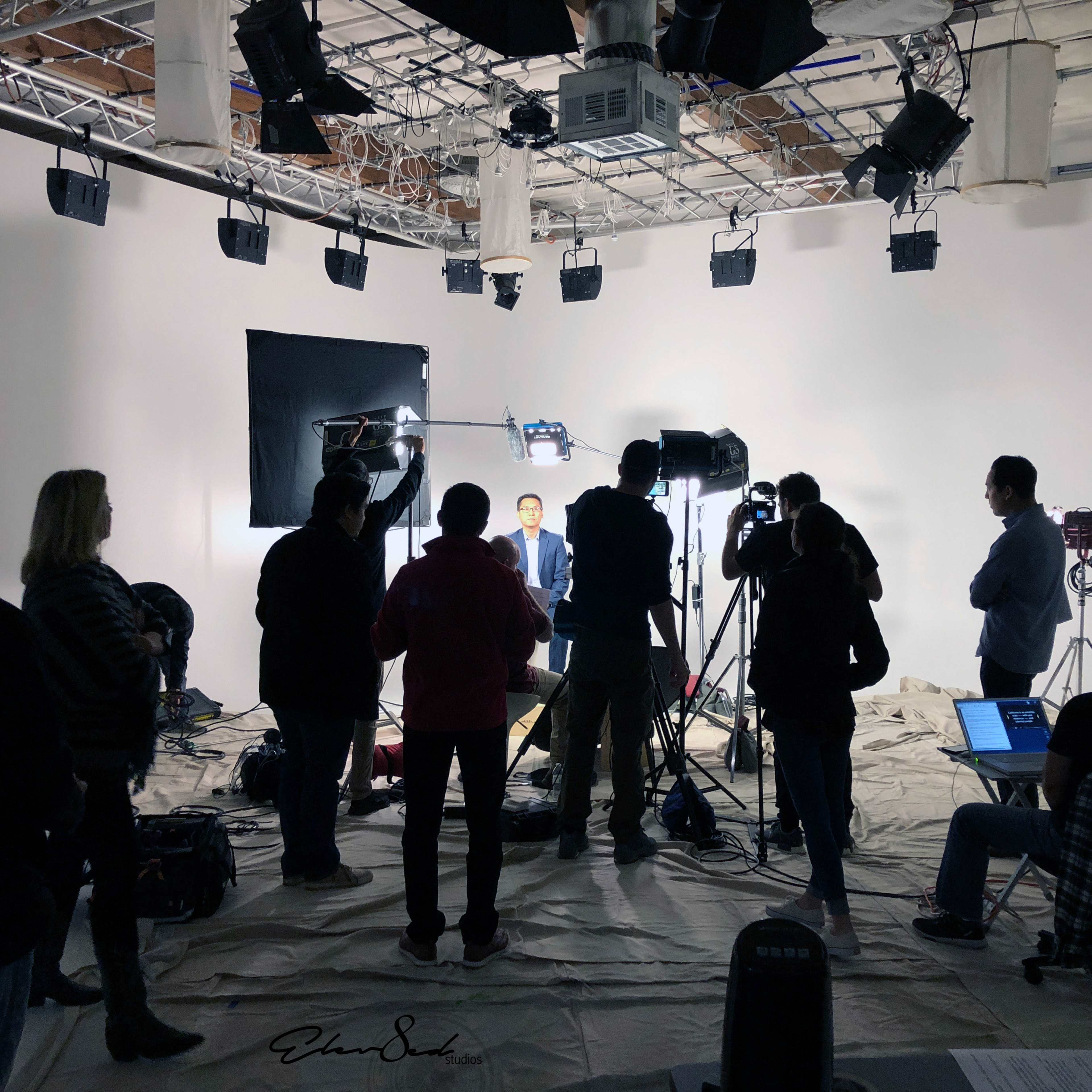 A group of people operates cameras and lighting equipment in a studio setting, focusing on a seated individual against a white backdrop.