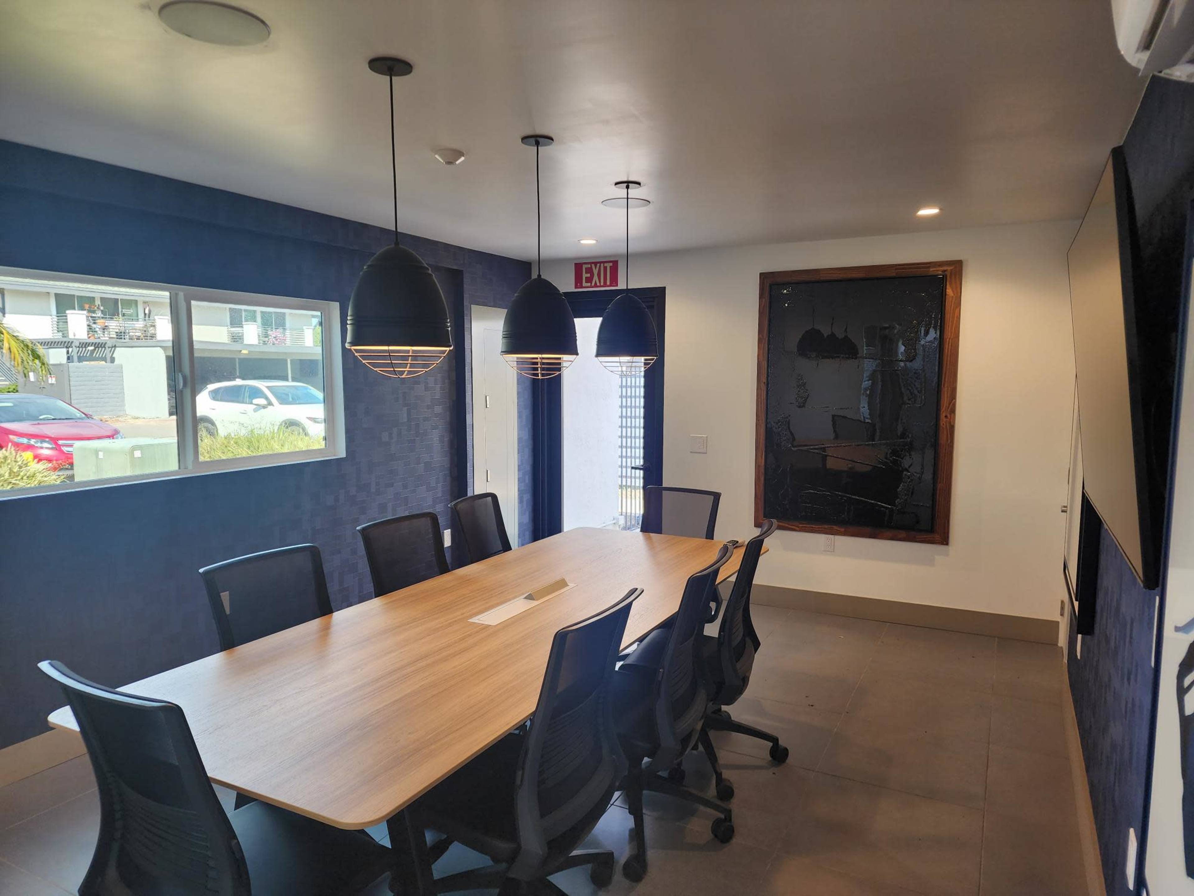 A conference room features a long wooden table surrounded by black chairs, with pendant lights overhead and a large framed artwork on the wall.