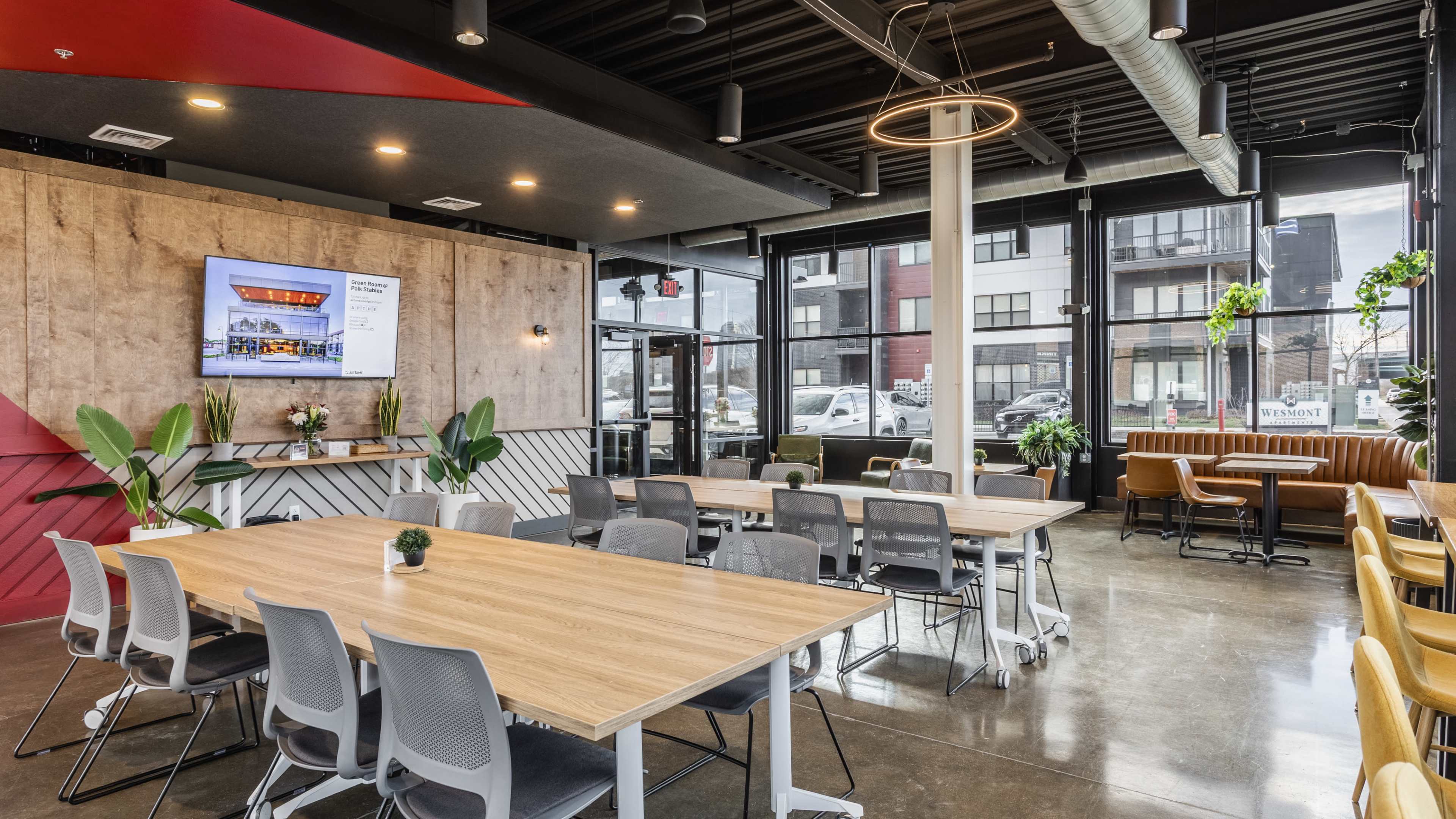 The image shows a modern indoor dining area featuring wooden tables, gray chairs, and large windows that allow natural light to enter.