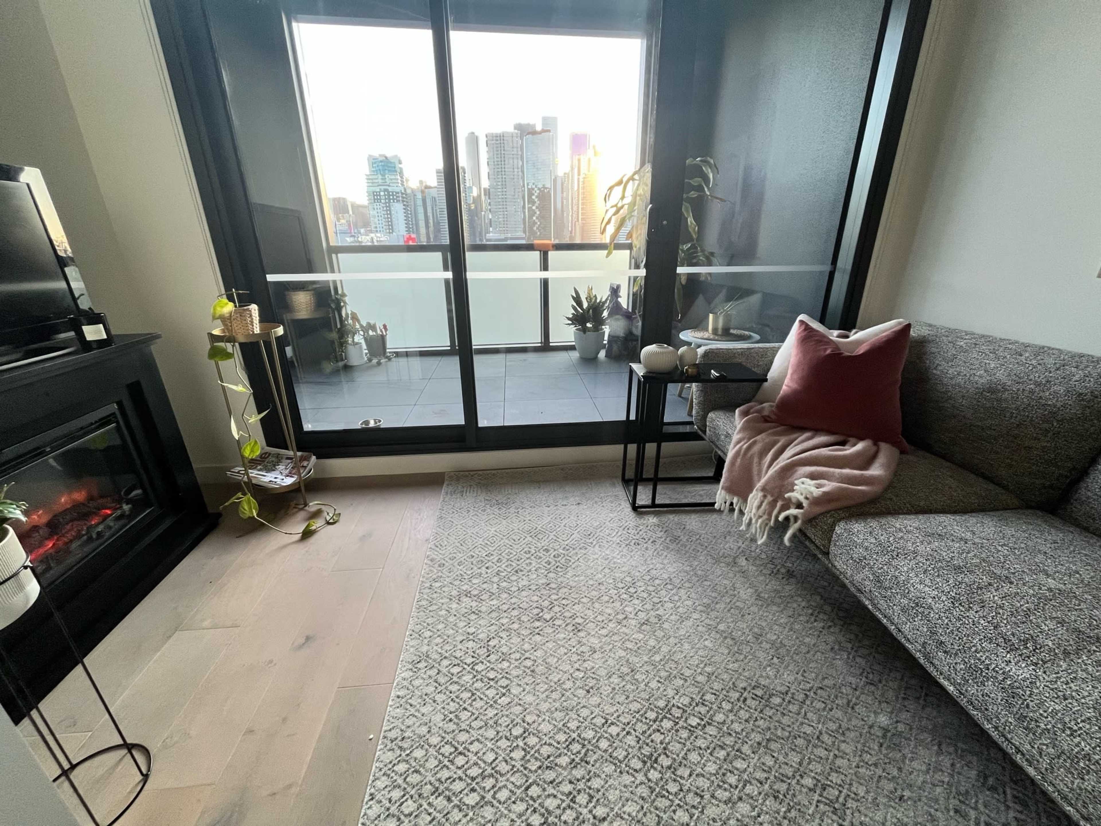 A cozy living room features a couch, a side table, and a view of a city skyline through large glass doors.