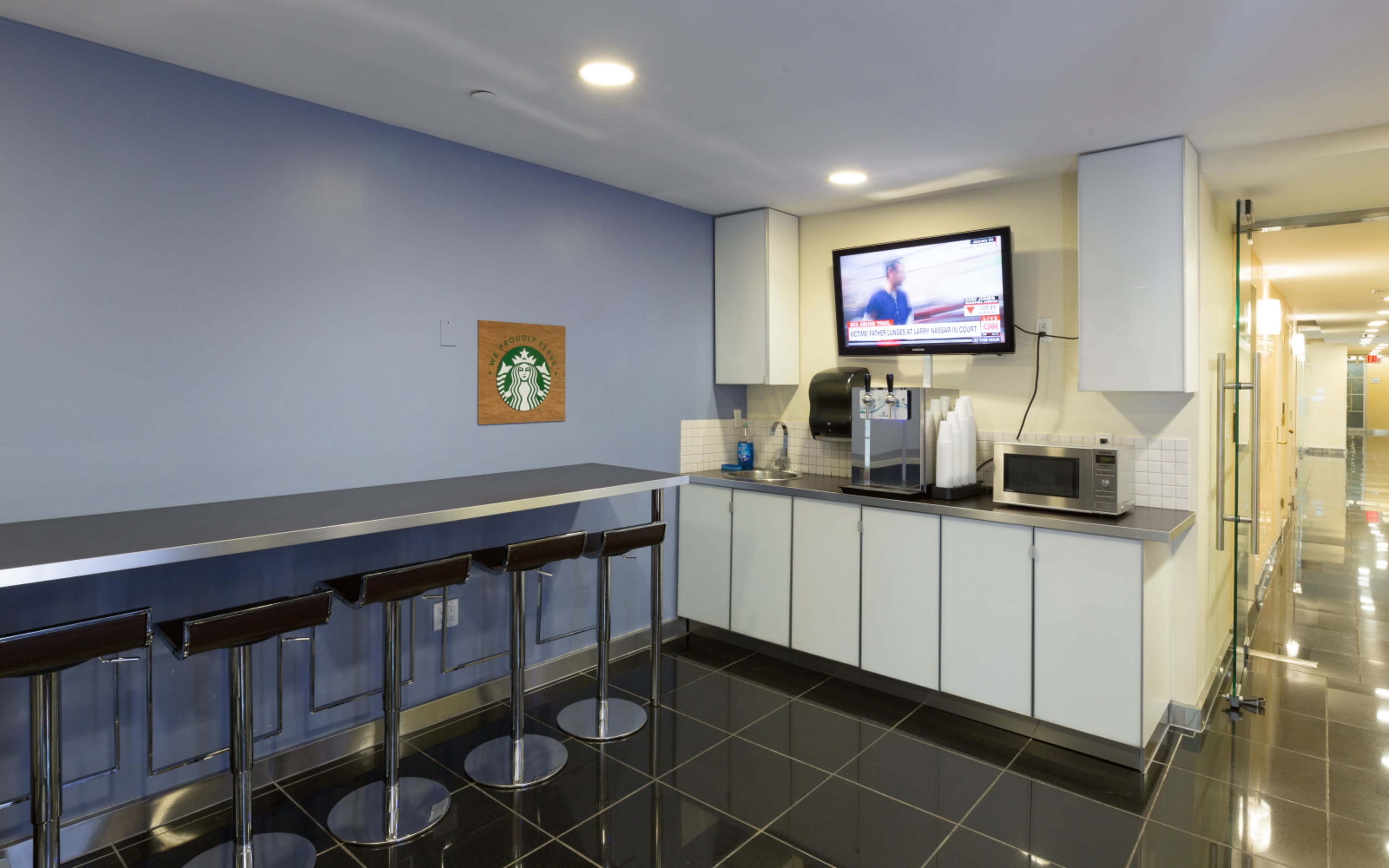 A modern kitchen area with a bar counter and stools, a microwave, and a television mounted on the wall displaying news.