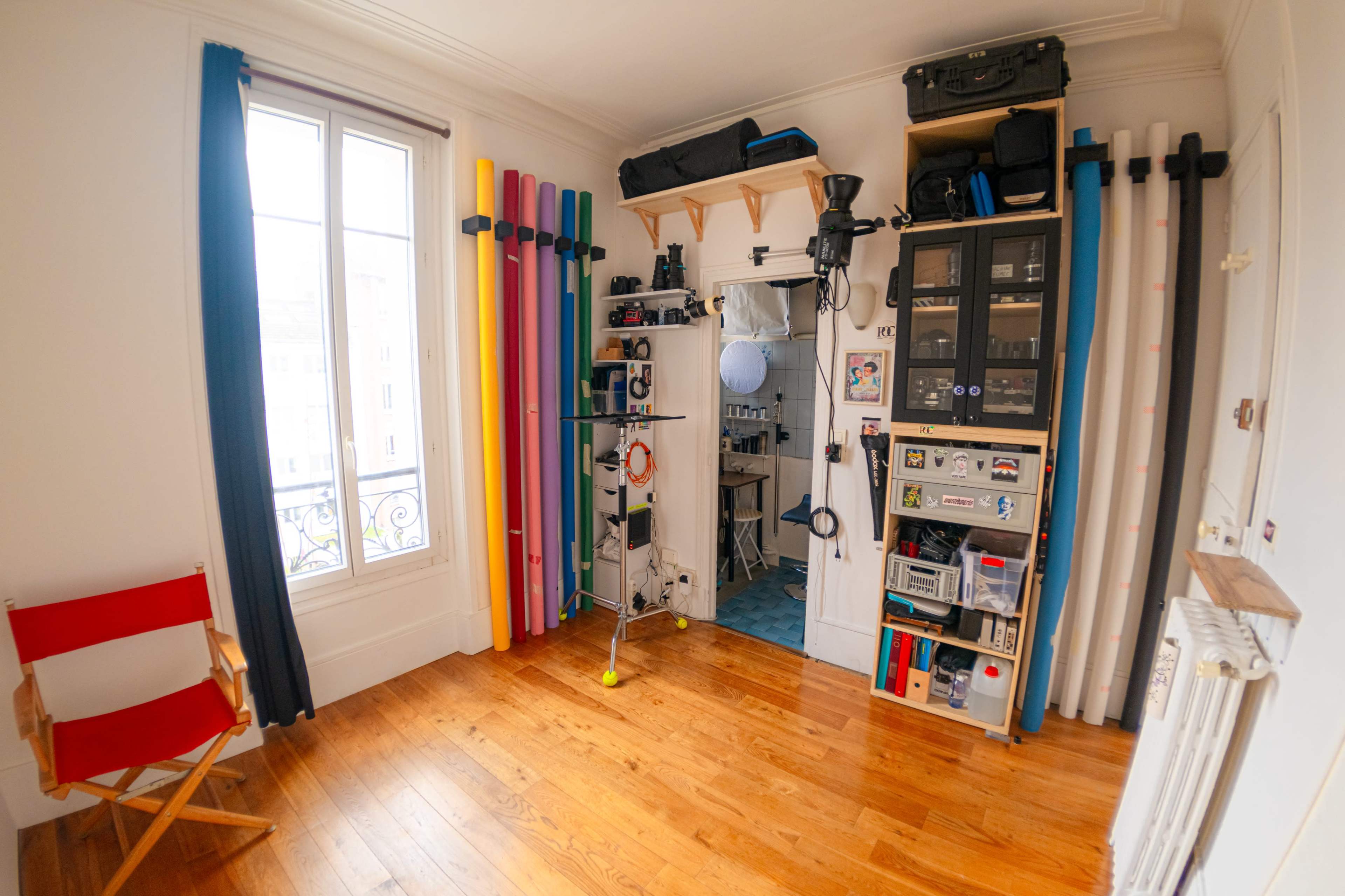 A small room features wooden flooring, a red director's chair, various colored poles arranged against the wall, and shelves with storage boxes.