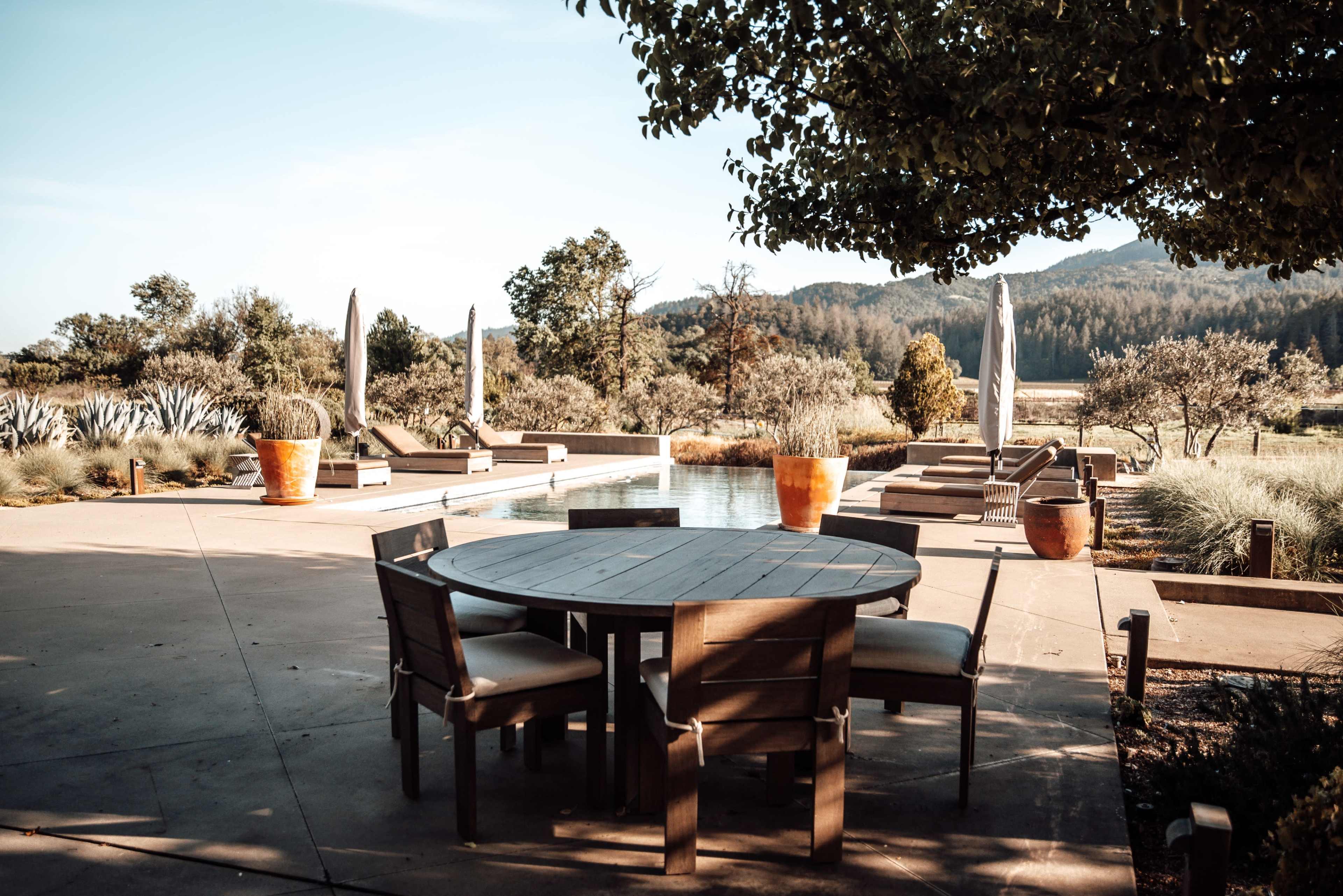 A wooden table with chairs is set near a swimming pool surrounded by landscaped gardens and lounge chairs.