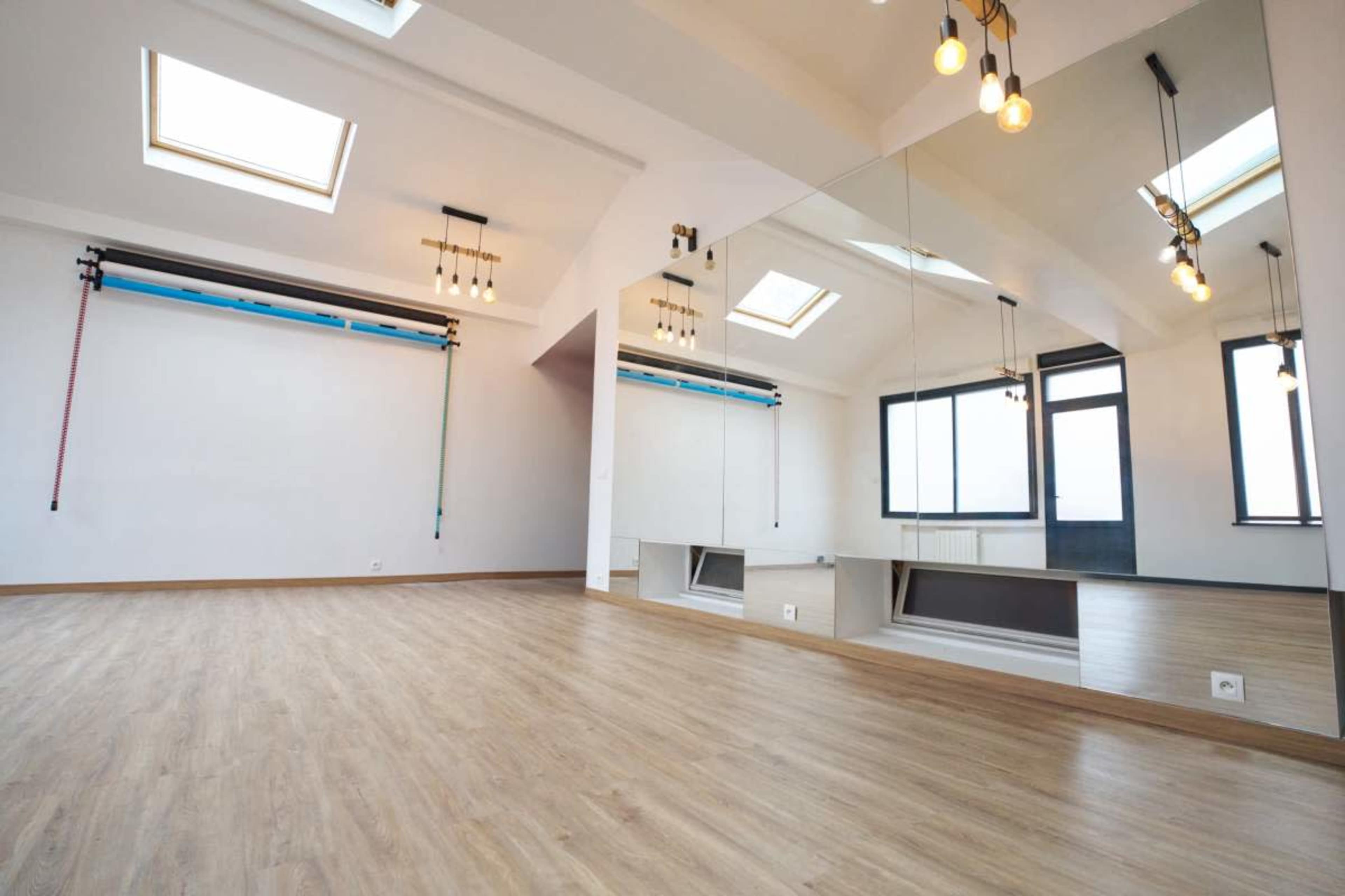 The image shows a spacious, well-lit room with a mirrored wall, large windows, and wooden flooring.