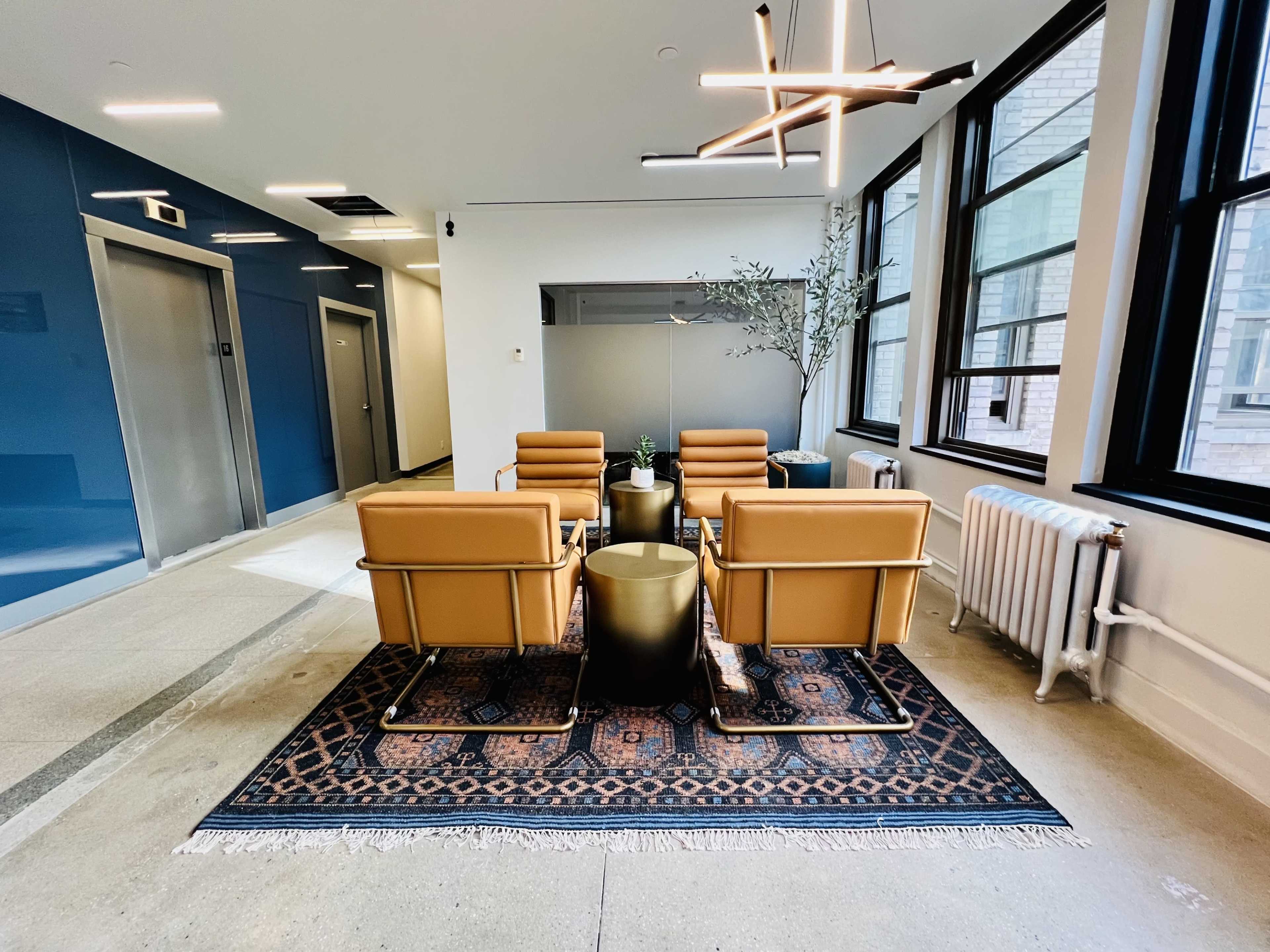 A modern waiting area with four tan chairs arranged around a small round table on a patterned rug, accompanied by a potted plant and large windows.