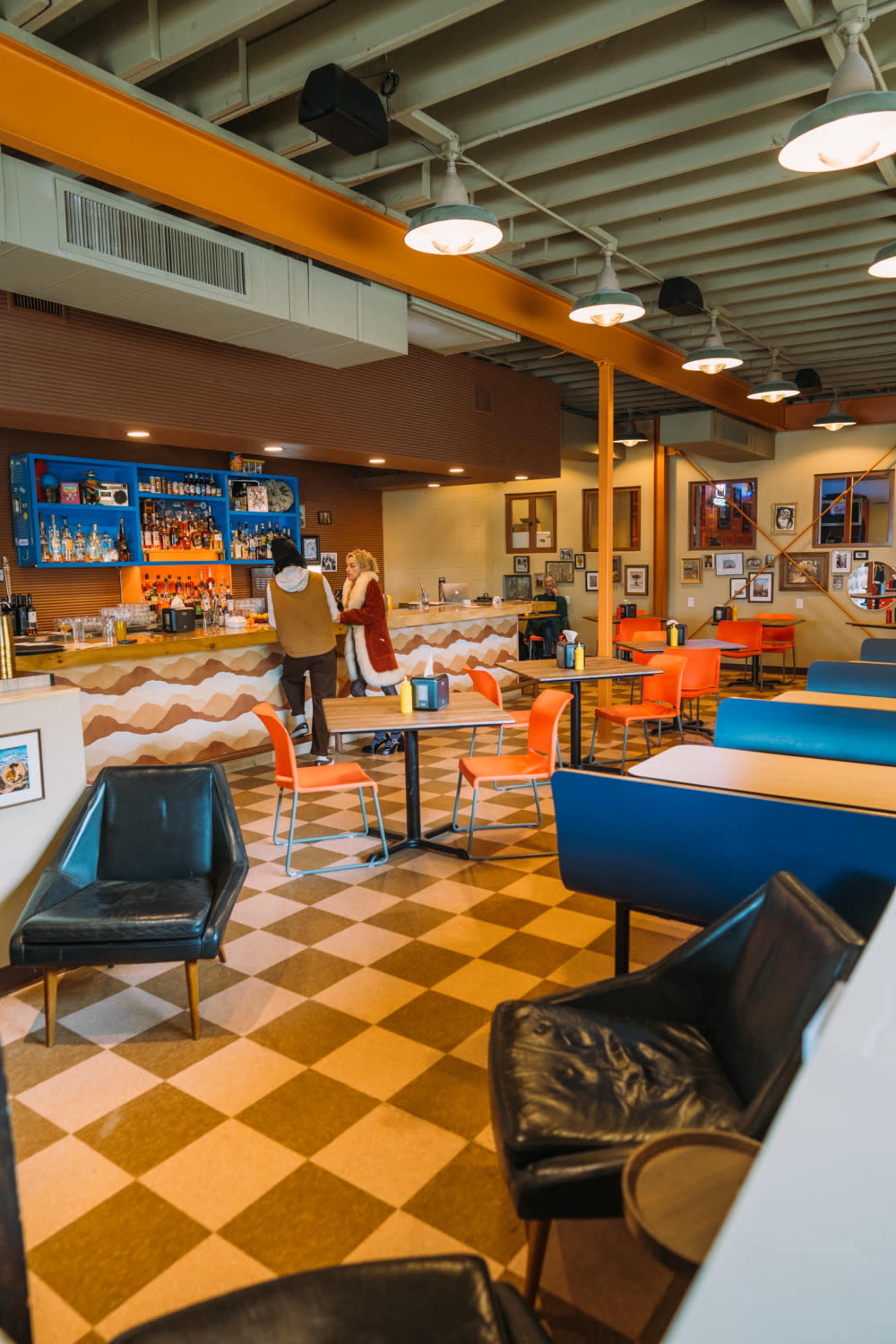 A spacious diner interior features a mix of colorful chairs at tables, a service counter in the background, and two people interacting near the bar area.
