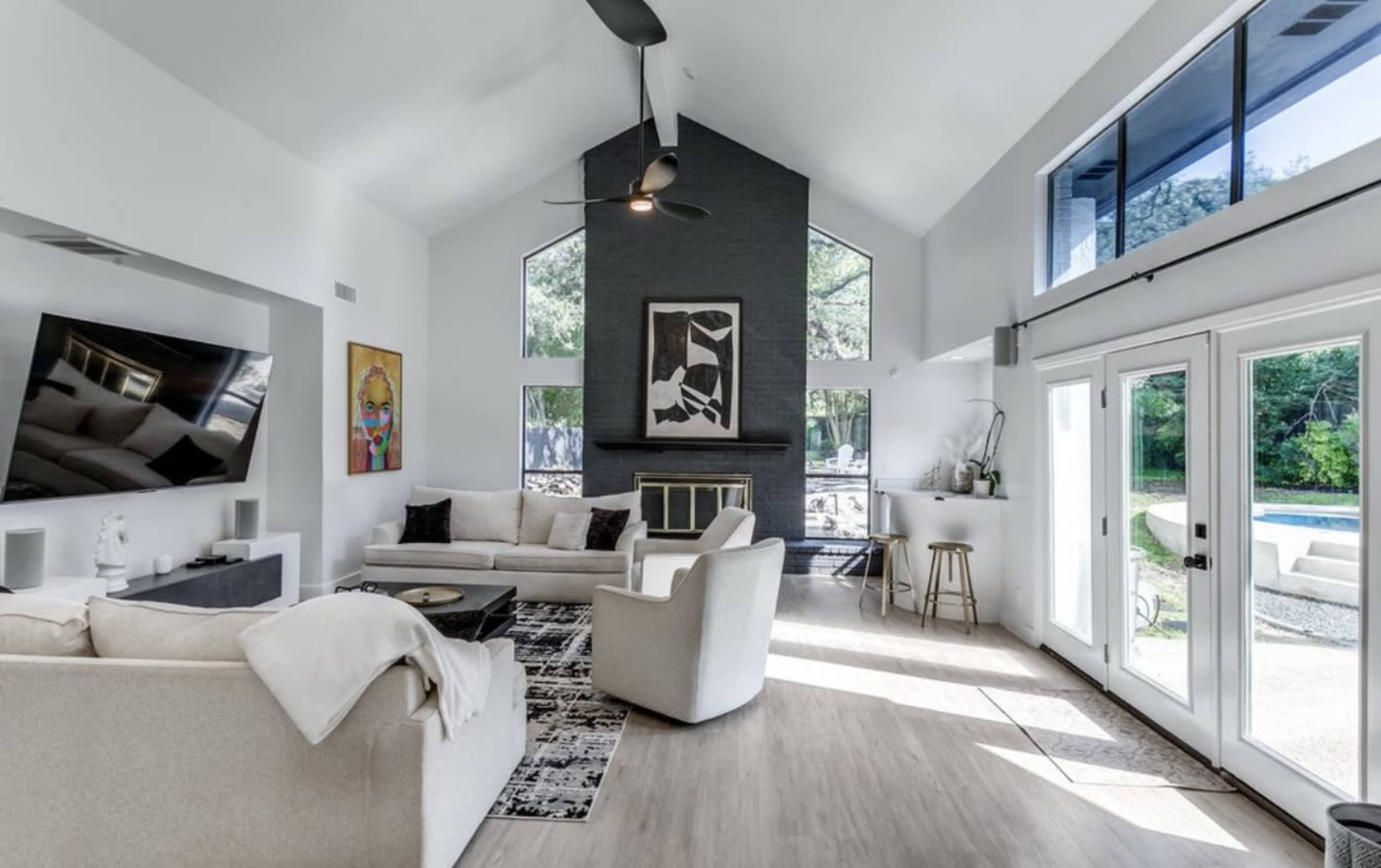 A spacious living room features a high ceiling, large windows, a modern fireplace, and contemporary furniture arranged around a patterned rug.