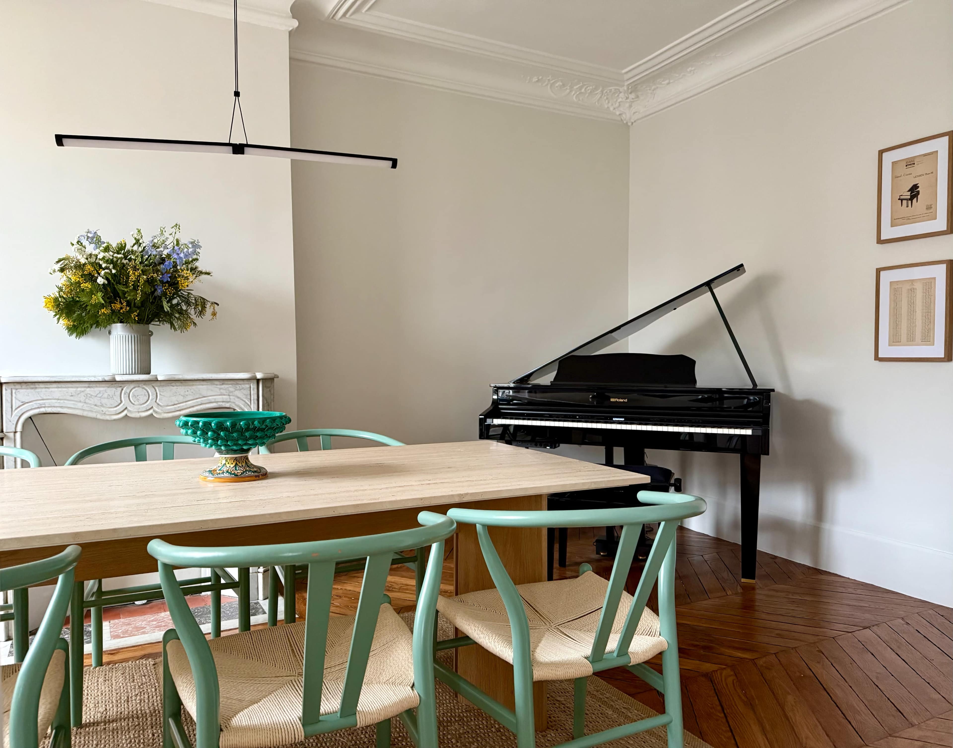 A dining area features a long wooden table surrounded by turquoise chairs, with a grand piano in one corner and a vase of flowers on a side table.