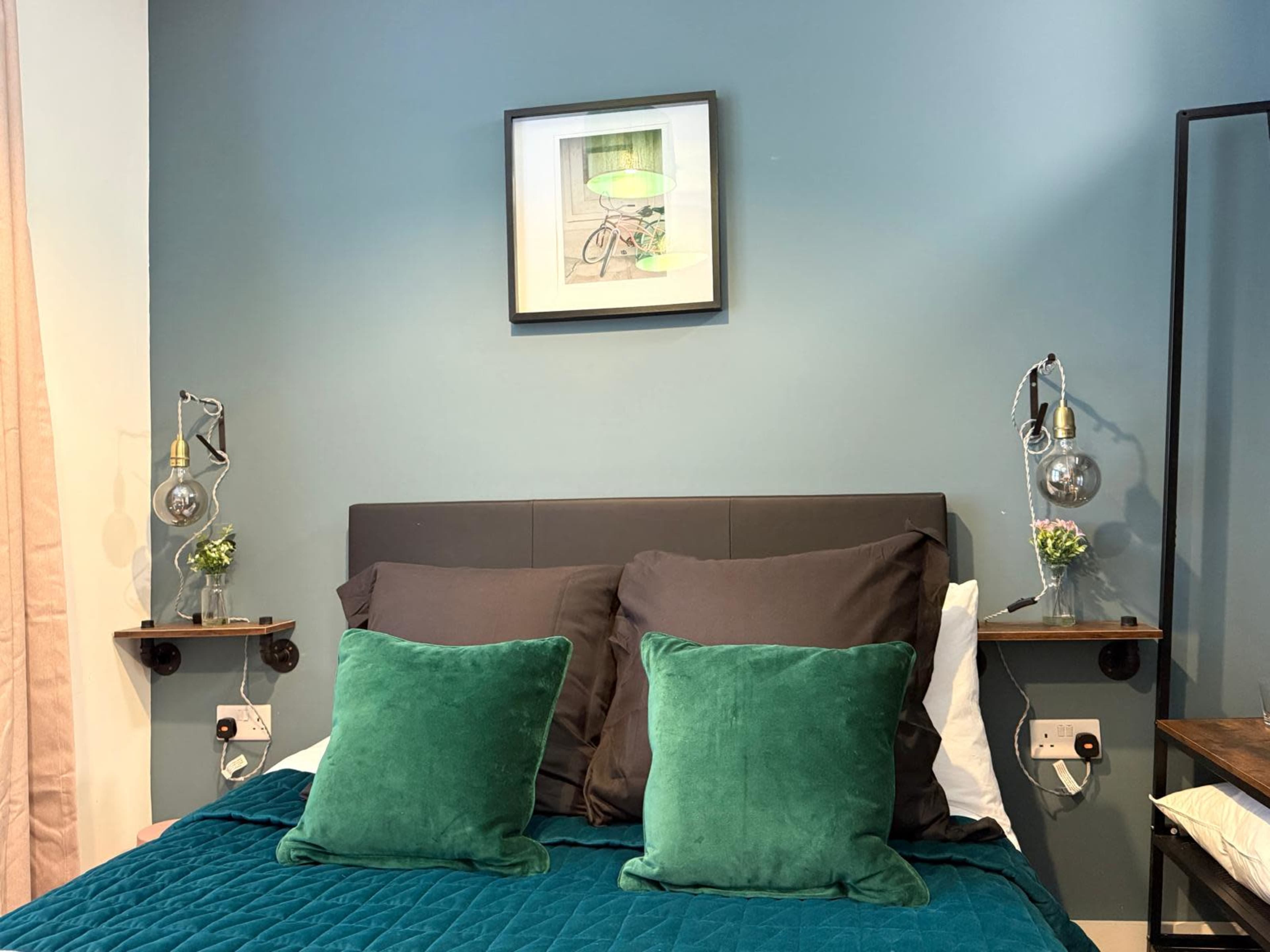The image shows a neatly made bed with dark pillows and green cushions against a blue wall, accompanied by framed artwork and decorative lamps on either side.