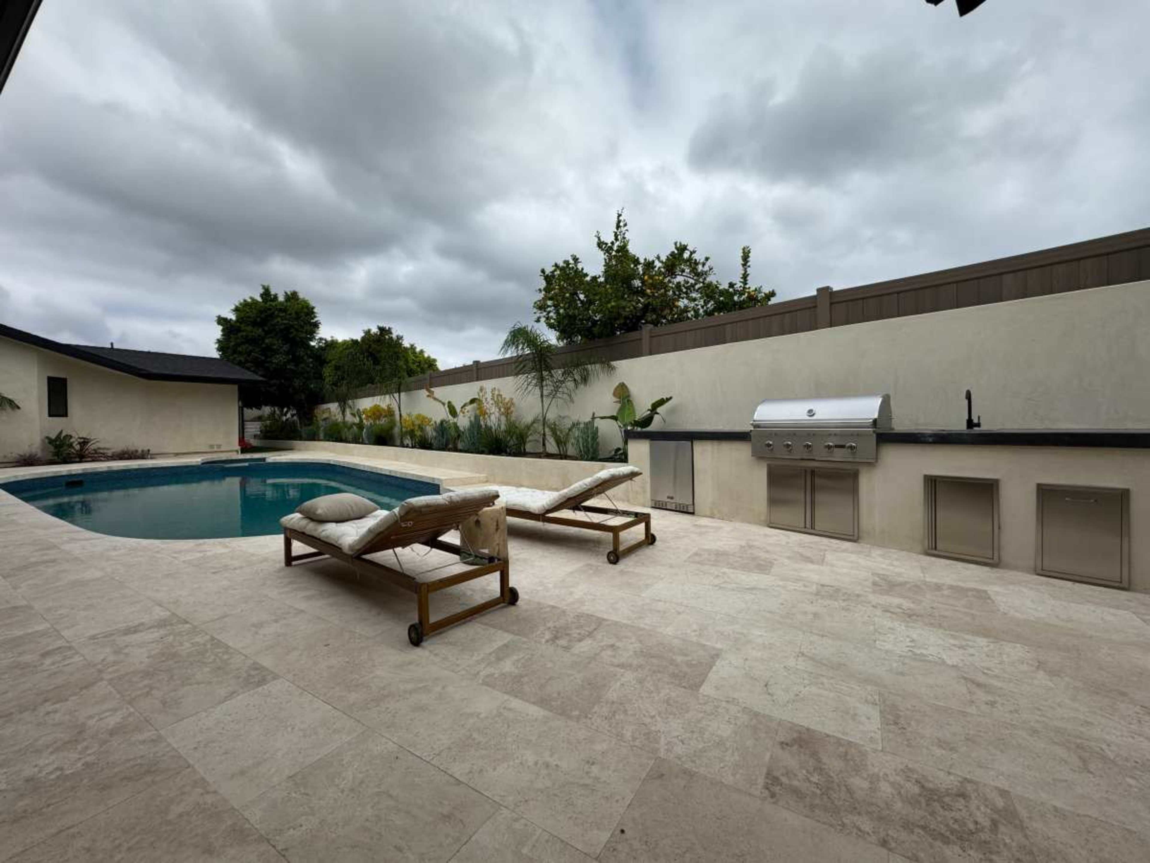 The image shows a backyard with a swimming pool, two lounging chairs, and a barbecue area surrounded by a low wall and greenery.