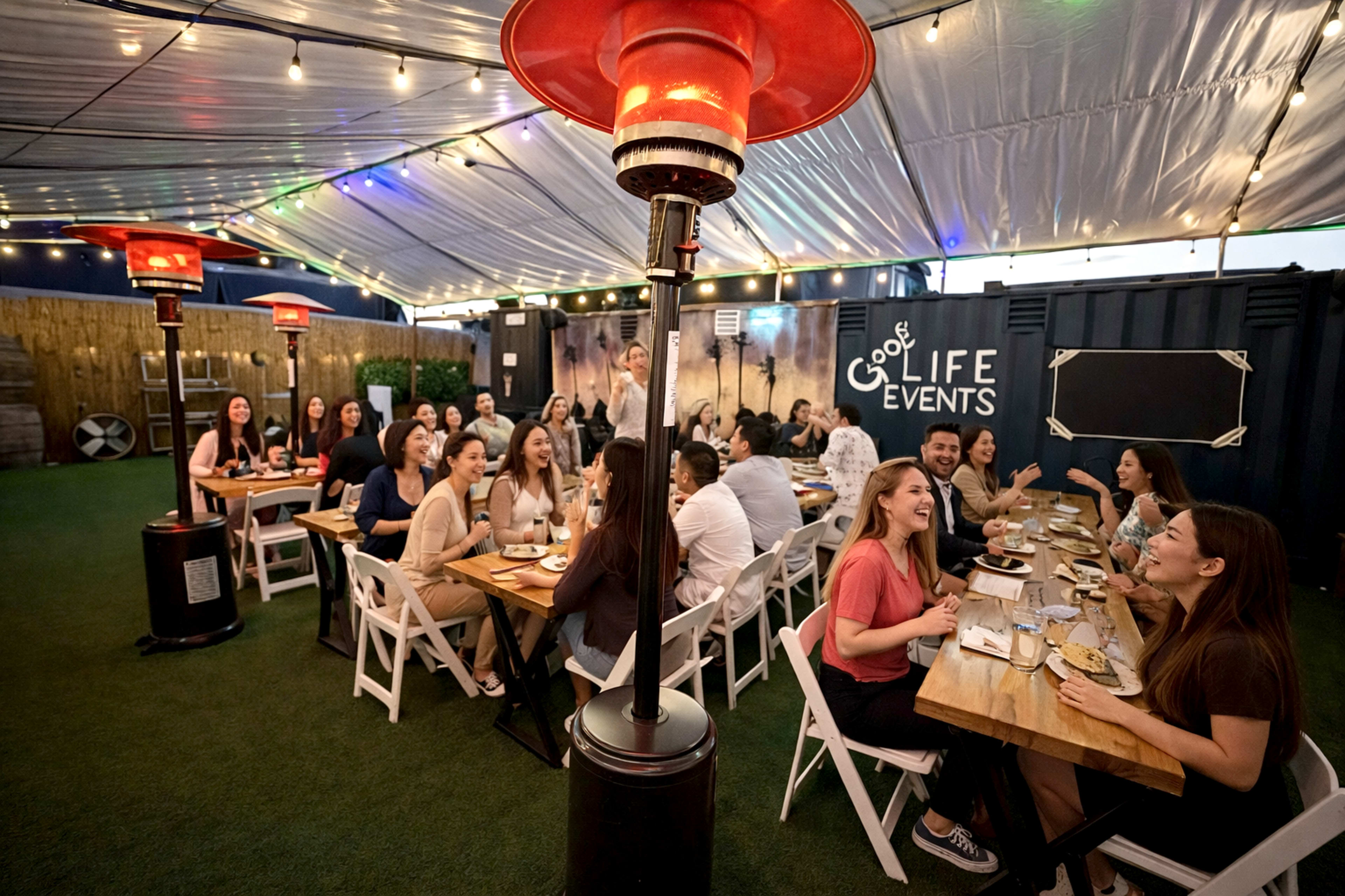 A group of people sits at long tables under a tent, enjoying a meal in a festive outdoor setting with string lights.