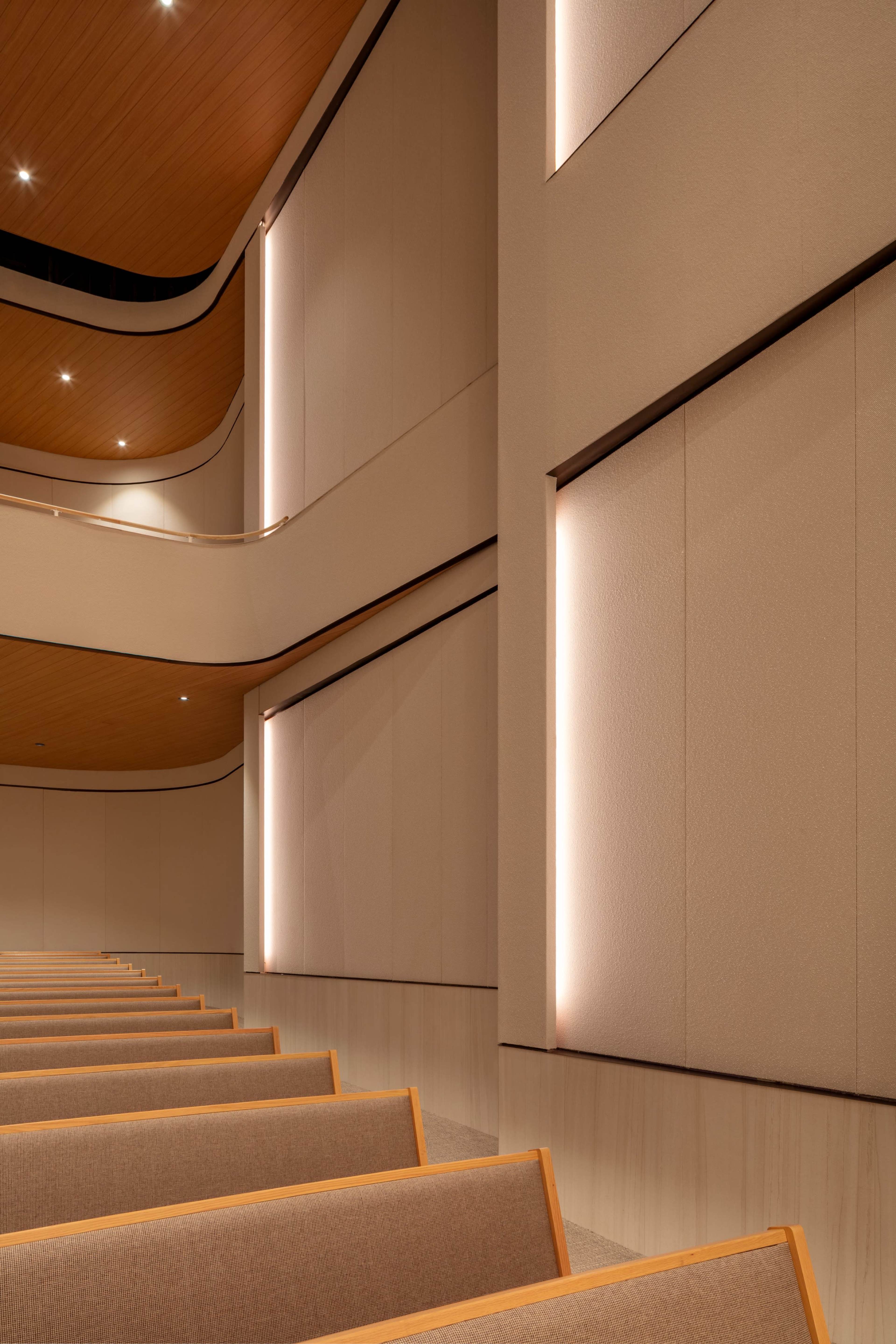 The image shows a modern auditorium with rows of wooden pews facing a softly lit interior featuring textured walls and curved ceilings.